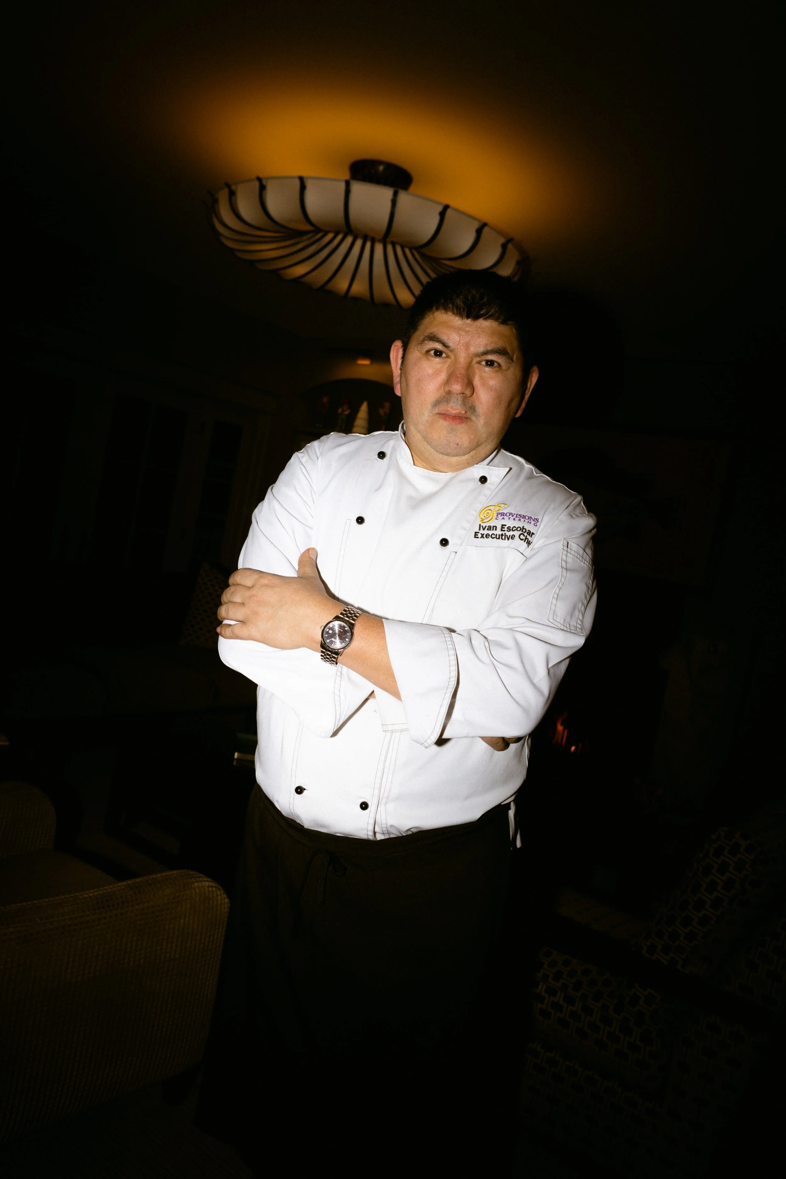 A male chef in a white chef's coat with black buttons and a watch, standing with arms crossed in a dimly lit room with a decorative ceiling fixture.