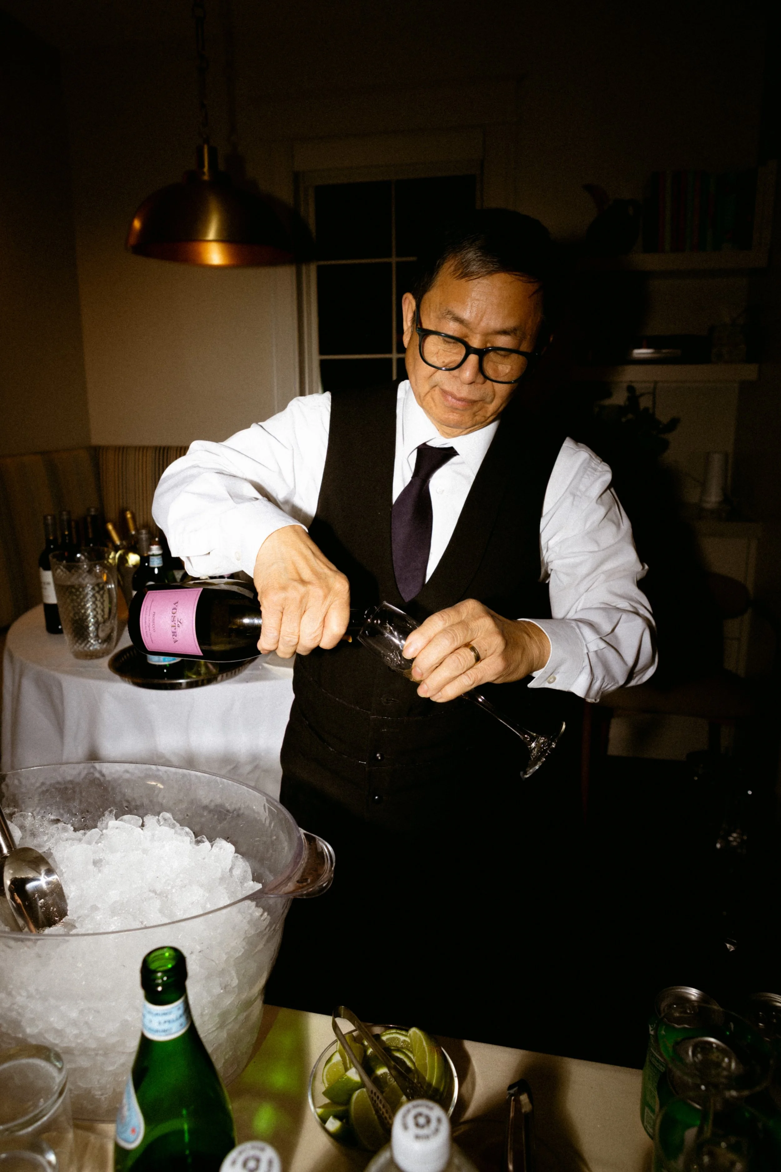 A man with glasses and a black vest pours a drink from a bottle into a glass at a dinner party, with bottles and lime slices on the table.