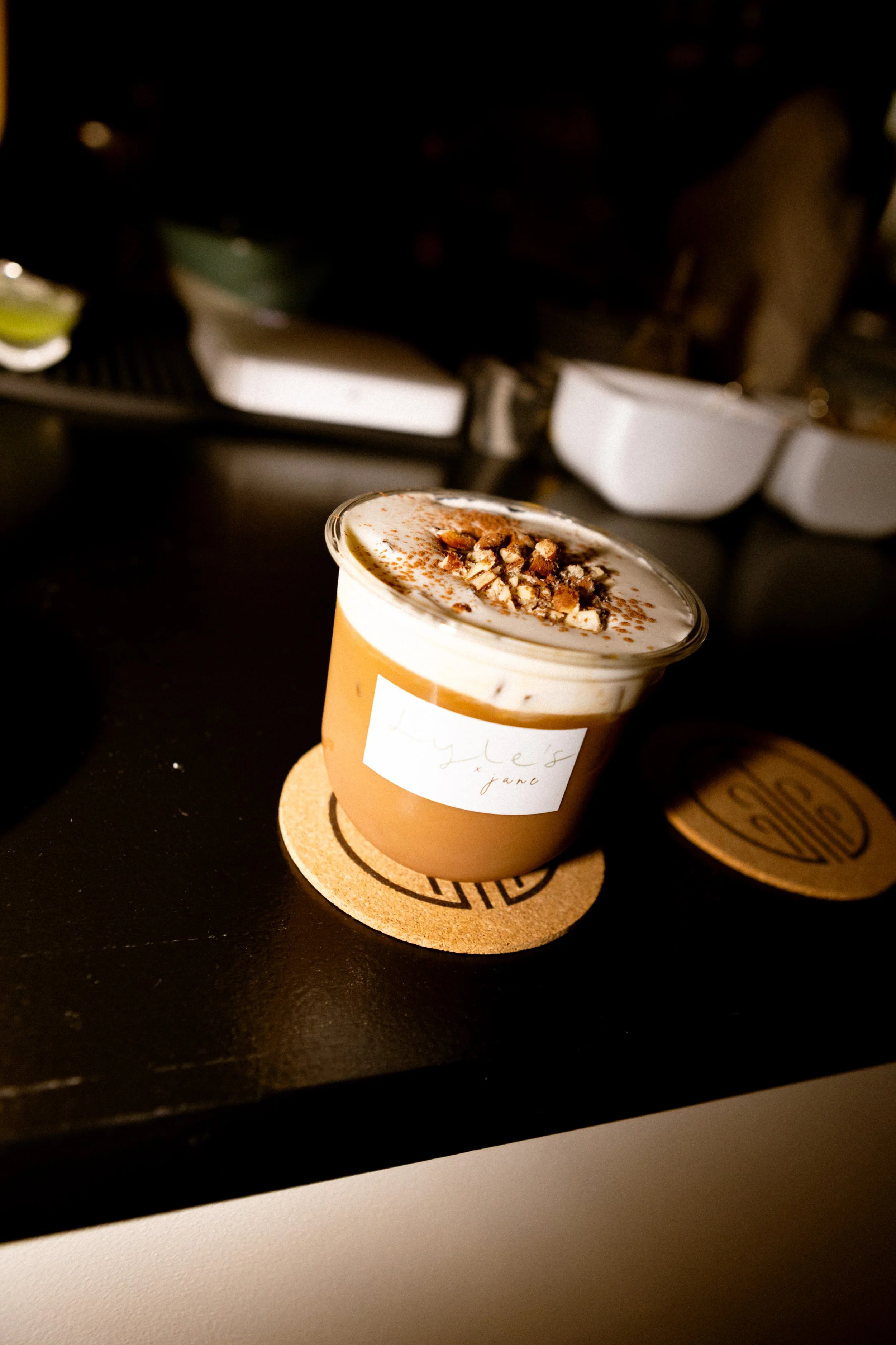 A coffee cup with whipped cream and chopped nuts on top, placed on a cork coaster on a black surface.