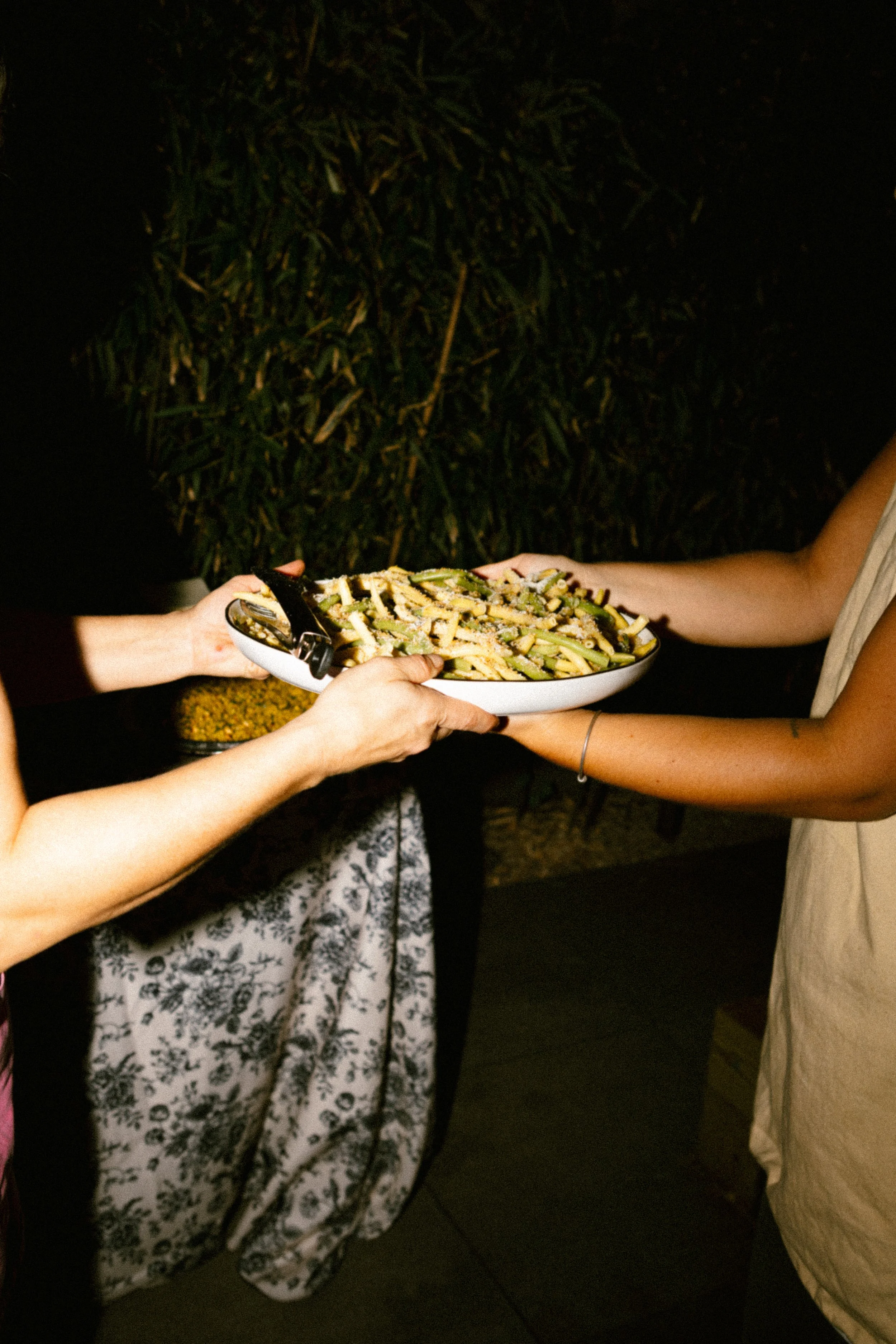 Two people exchanging a plate of pasta at night outside.