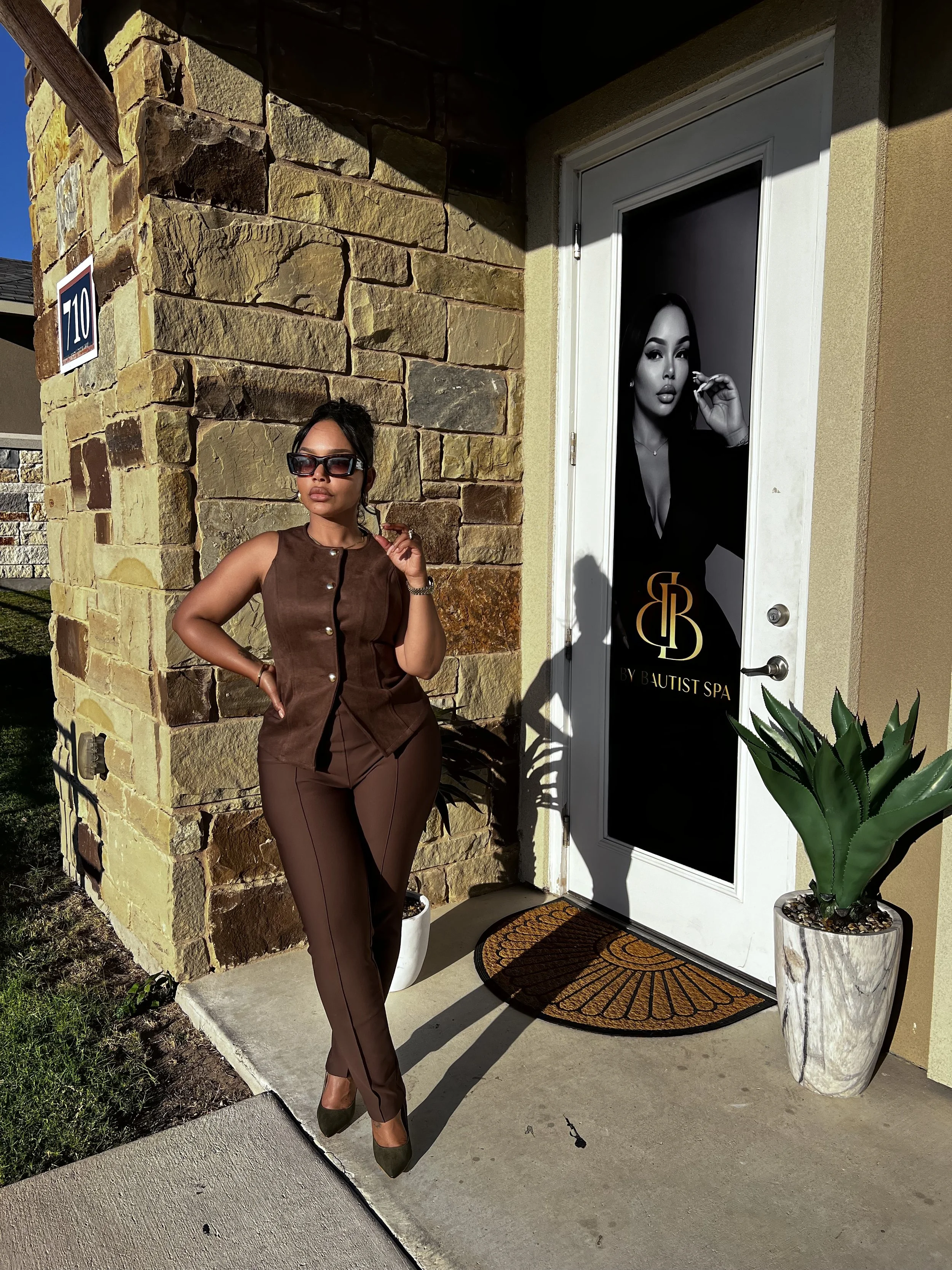 Woman dressed in brown standing outside a spa with a door featuring an advertisement with a woman in black. She is wearing sunglasses and high heels, posing with one hand on her hip and the other near her face.