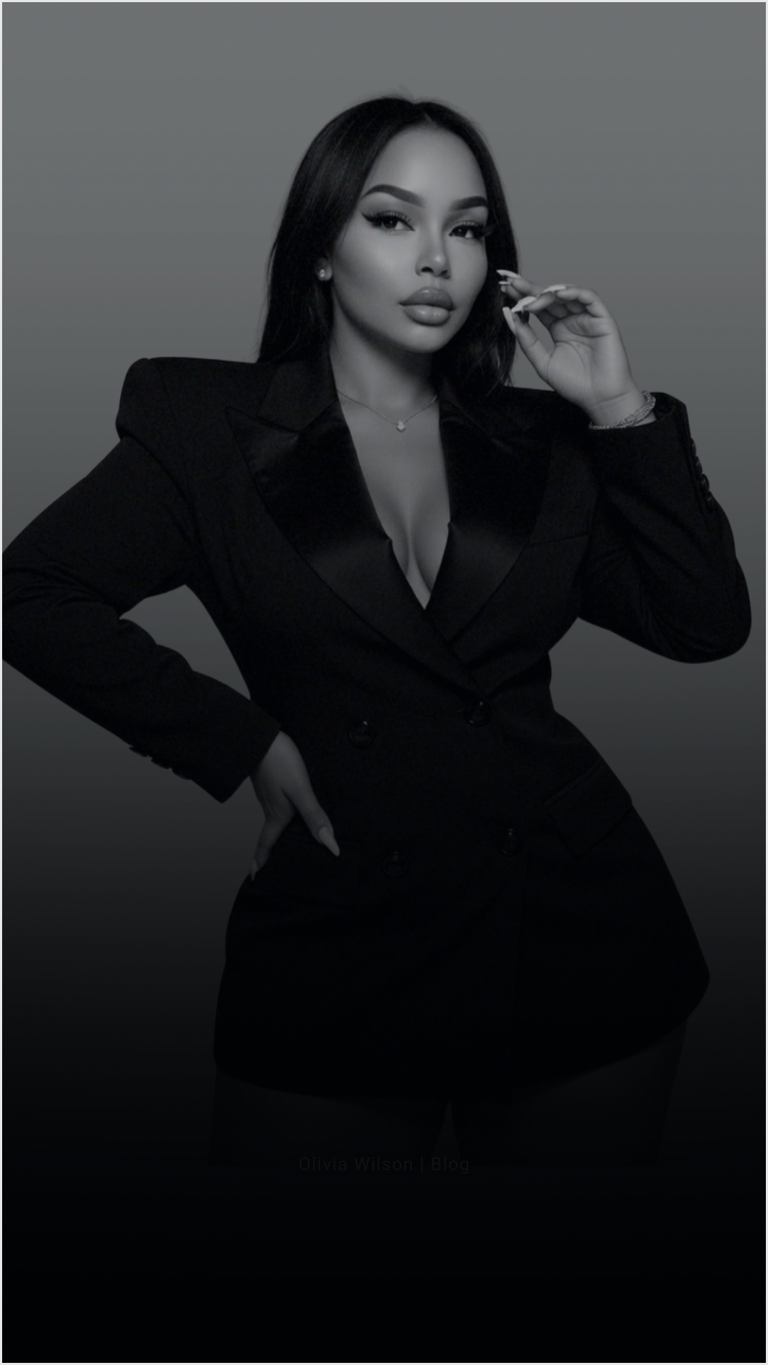 Black and white photograph of a woman wearing a tailored blazer with a low neckline, standing with one hand on her hip and the other near her face, with a confident expression.