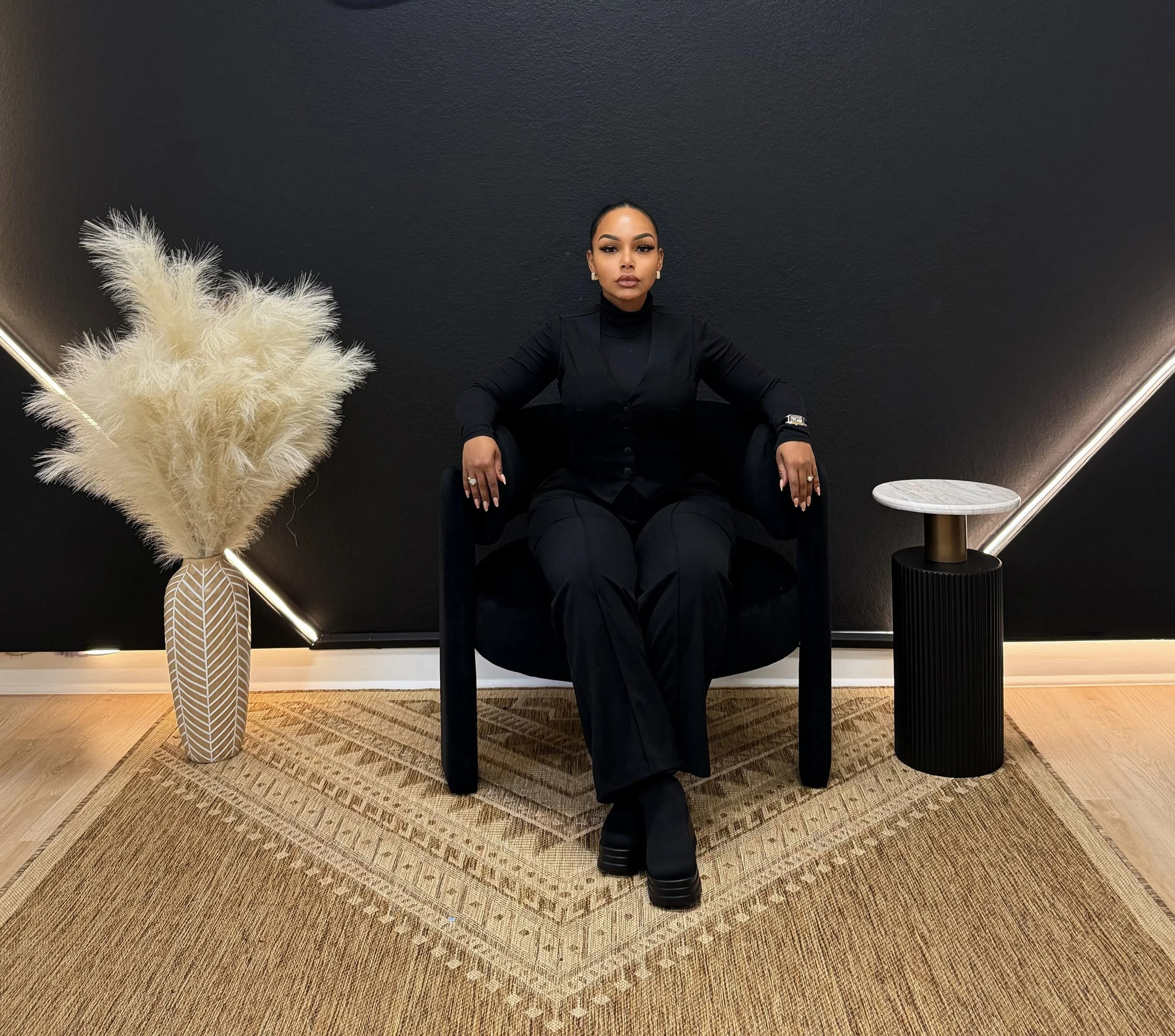 A woman dressed in black sitting on a black armchair with a black background, flanked by a white vase with pampas grass on her left and a small round side table on her right. The floor has a beige patterned rug.