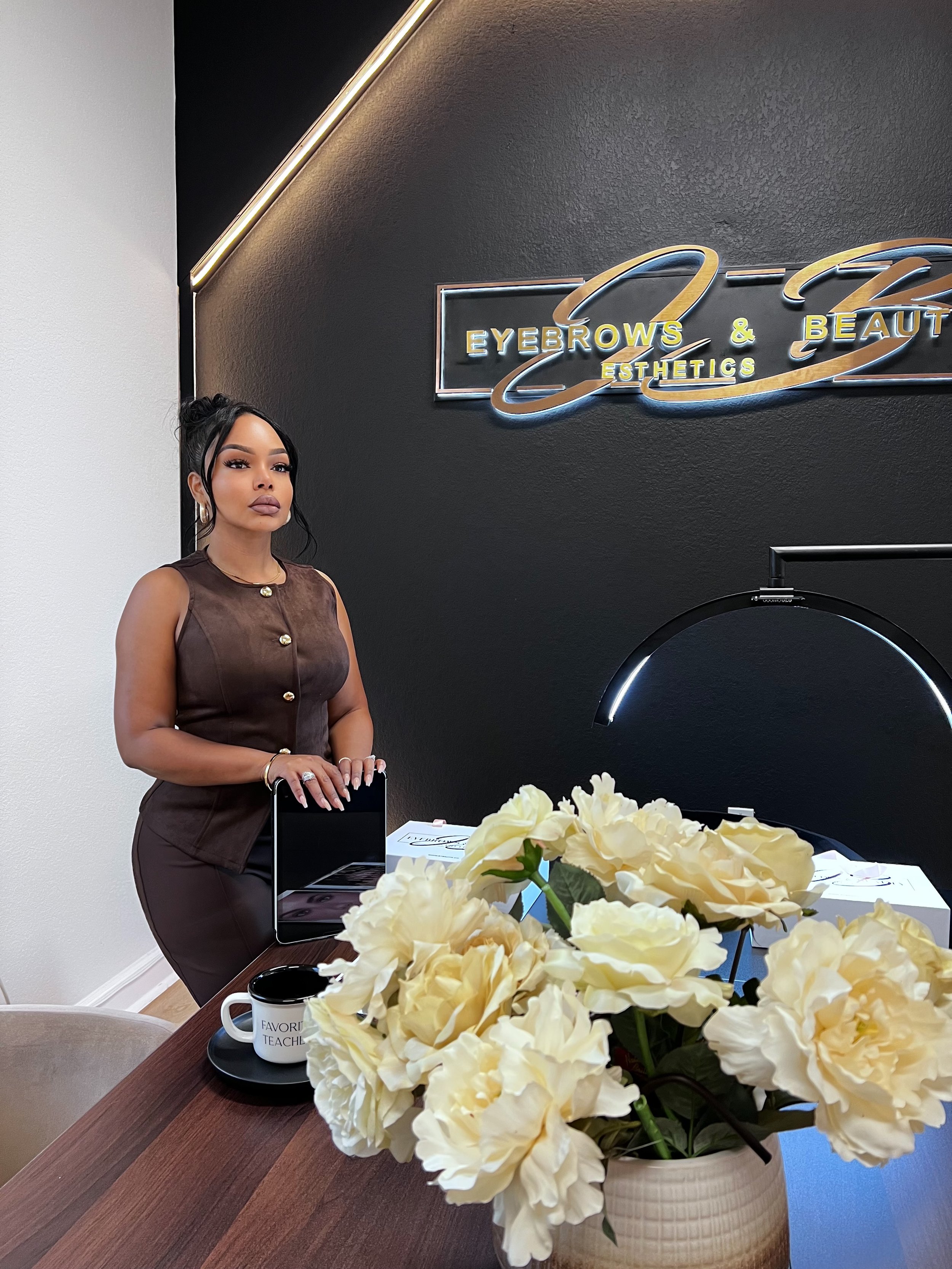 A woman with black hair styled in a bun, wearing a sleeveless brown dress, standing next to a table with a white flower arrangement in a beige vase, a coffee mug saying 'Favorite Teacher,' and a brochure or menu. She is in an office or beauty salon with a black wall featuring a neon sign that reads 'Eyebrows & Beauty Esthetics.'
