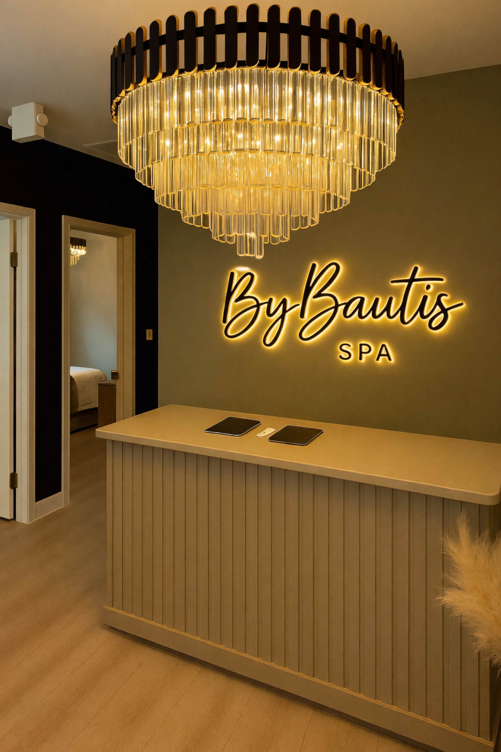 Interior of a spa reception area with a large chandelier, a sign that reads 'By Bautis SPA', and a wooden reception desk with two black tablets on top.