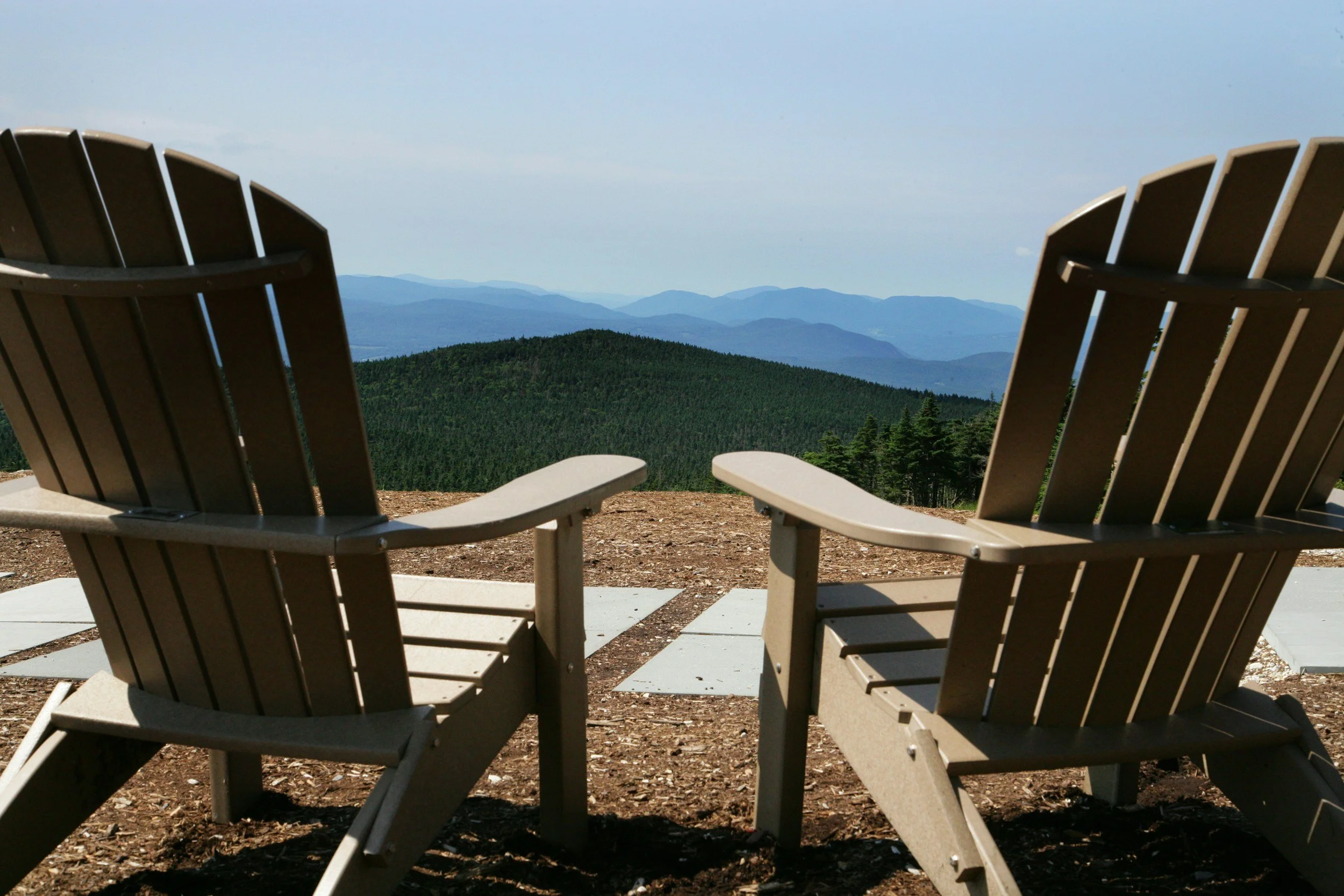 Two Adirondack chairs facing a scenic mountain view with green forests and blue mountains in the distance.
