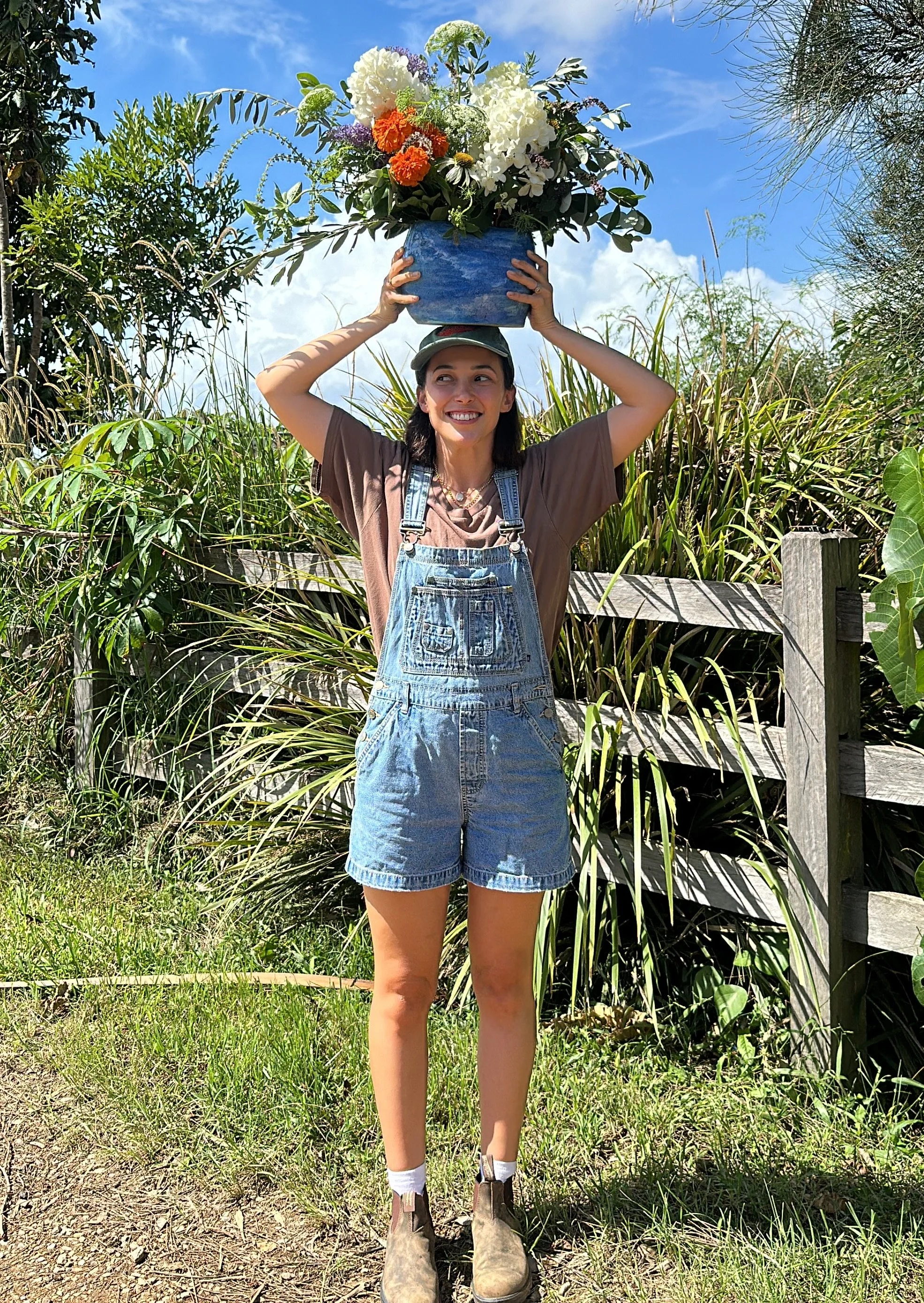 A woman in denim overalls, brown t-shirt, and a cap stands outdoors on grass, smiling, and holds a large blue pot filled with white, orange, purple, and green flowers above her head against a background of blue sky, green plants, and a wooden fence.