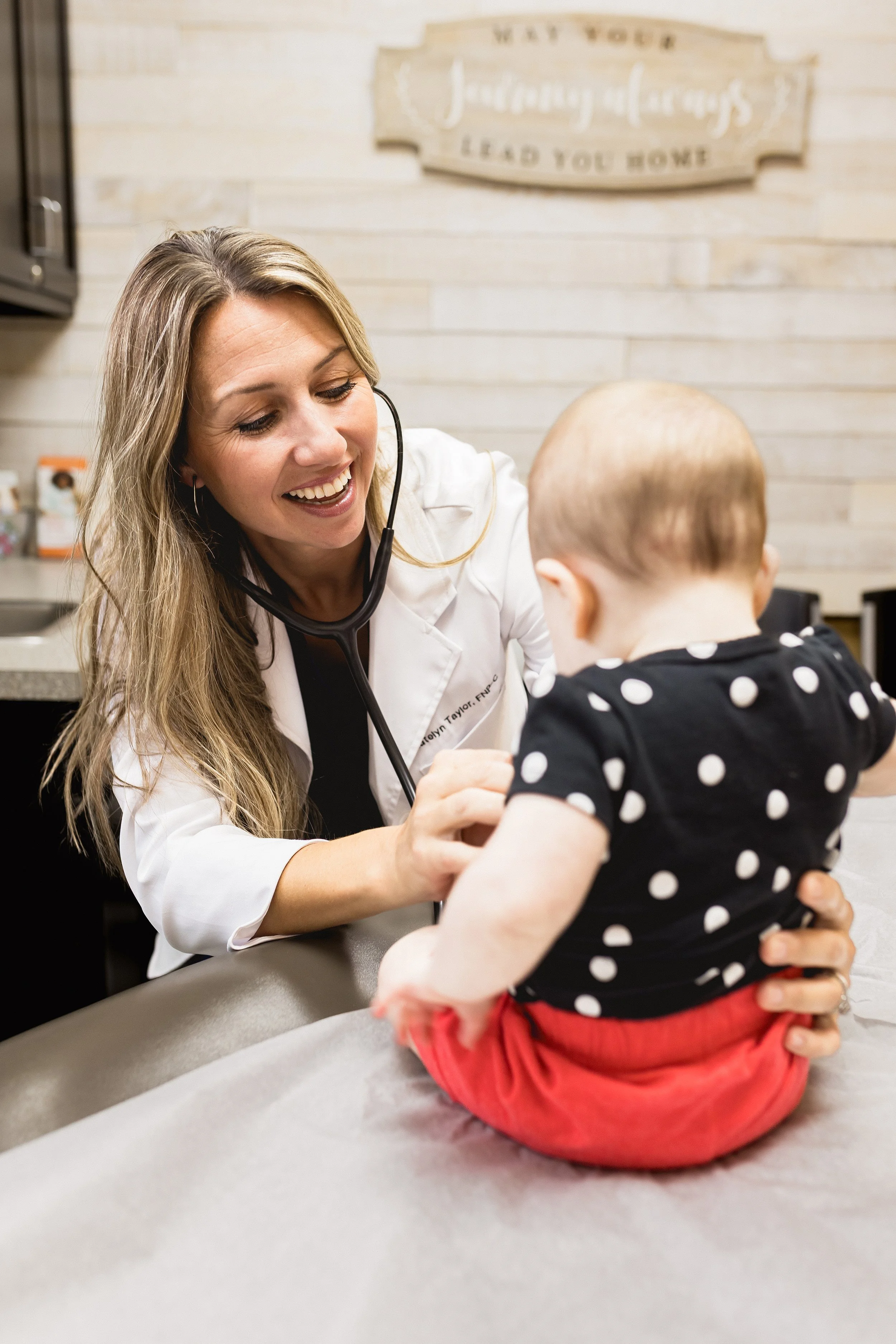 A female doctor wearing a white coat and stethoscope interacts with a young child sitting on an examination table in a medical office.