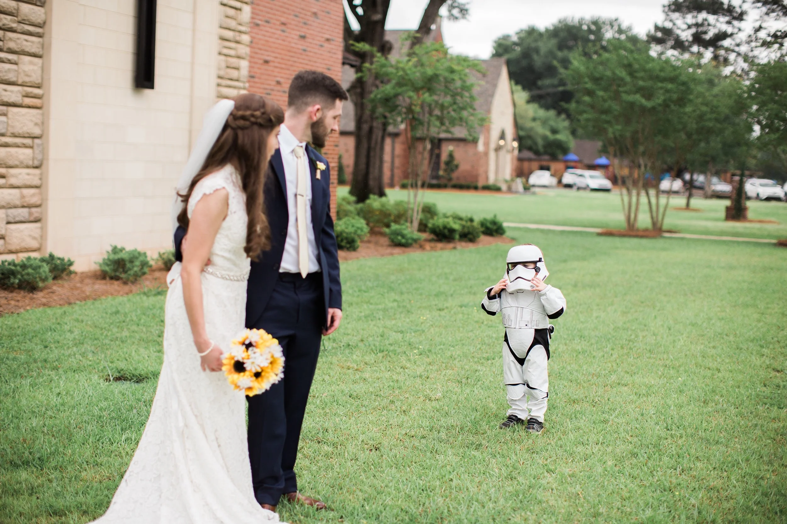A bride and groom stand outdoors on a grassy lawn, looking at a small child dressed as a Stormtrooper from Star Wars, who is holding a helmet over their face.