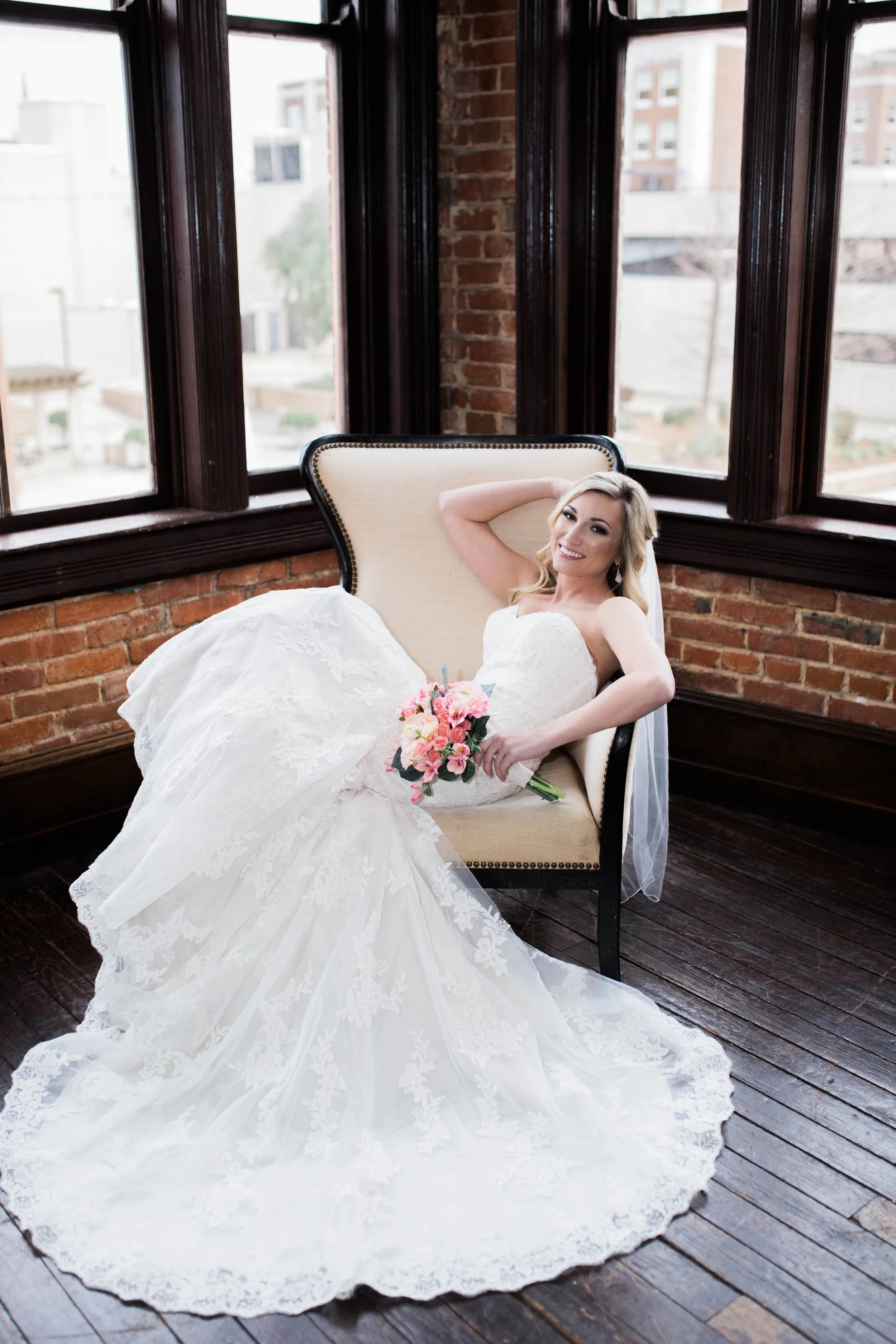 Bride in a white wedding dress sitting on a cream-colored armchair inside a room with brick walls and large windows, holding a pink bouquet, smiling at the camera.