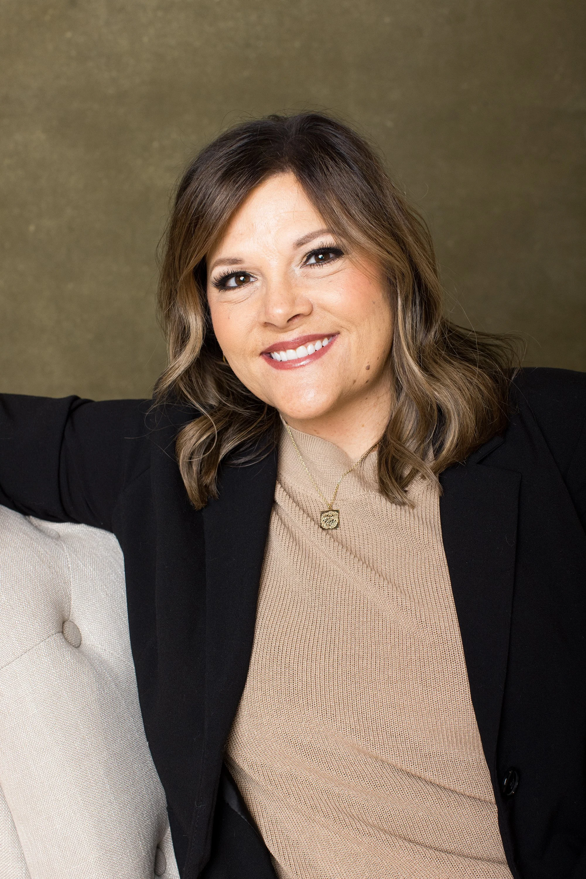 A woman with shoulder-length brown hair, wearing a black blazer and beige top, smiling and sitting on a light-colored sofa against a brown background.