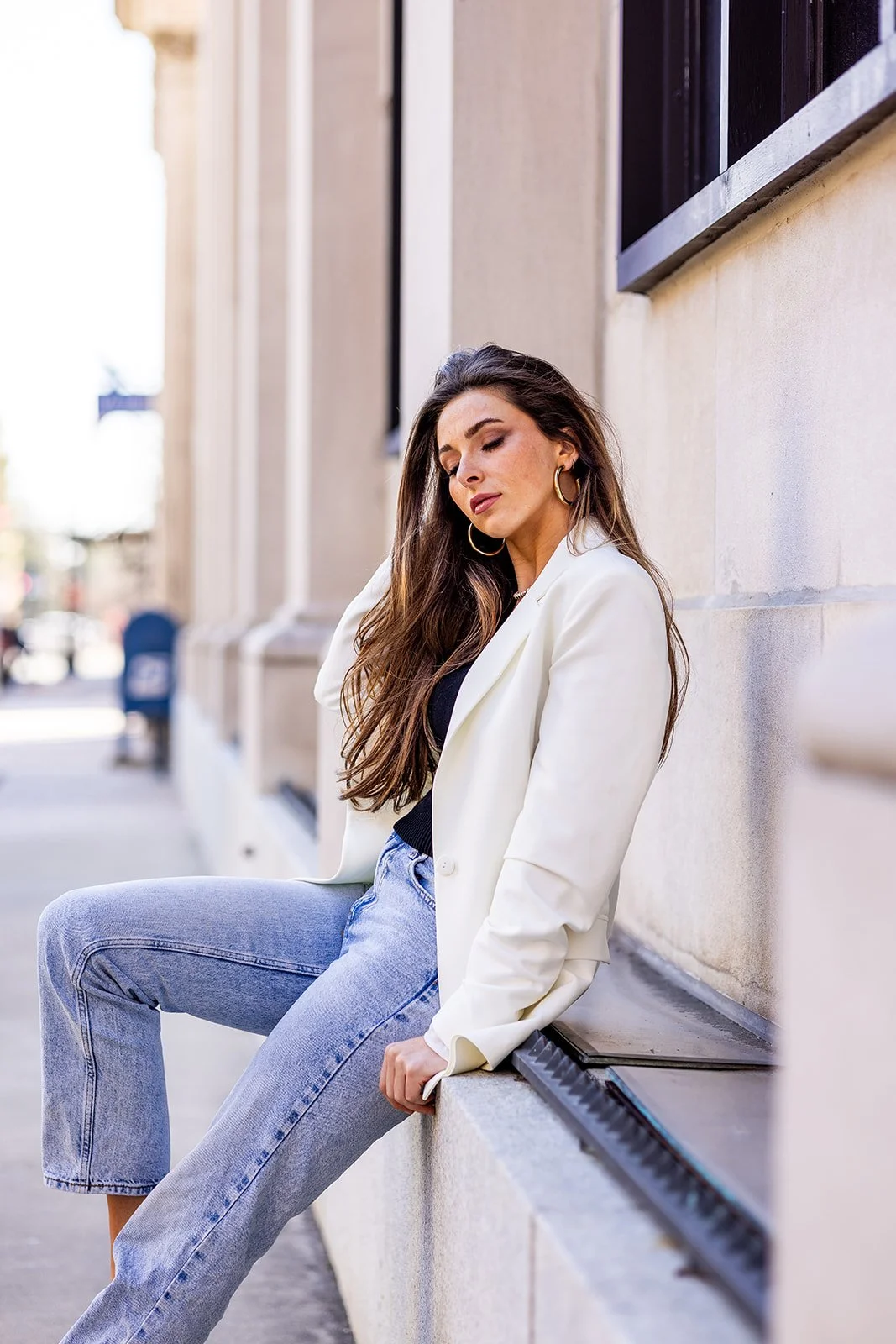 A woman with long brown hair sitting on a ledge against a building wall, wearing a white blazer, black top, light blue jeans, and gold hoop earrings, with her eyes closed and head tilted slightly.