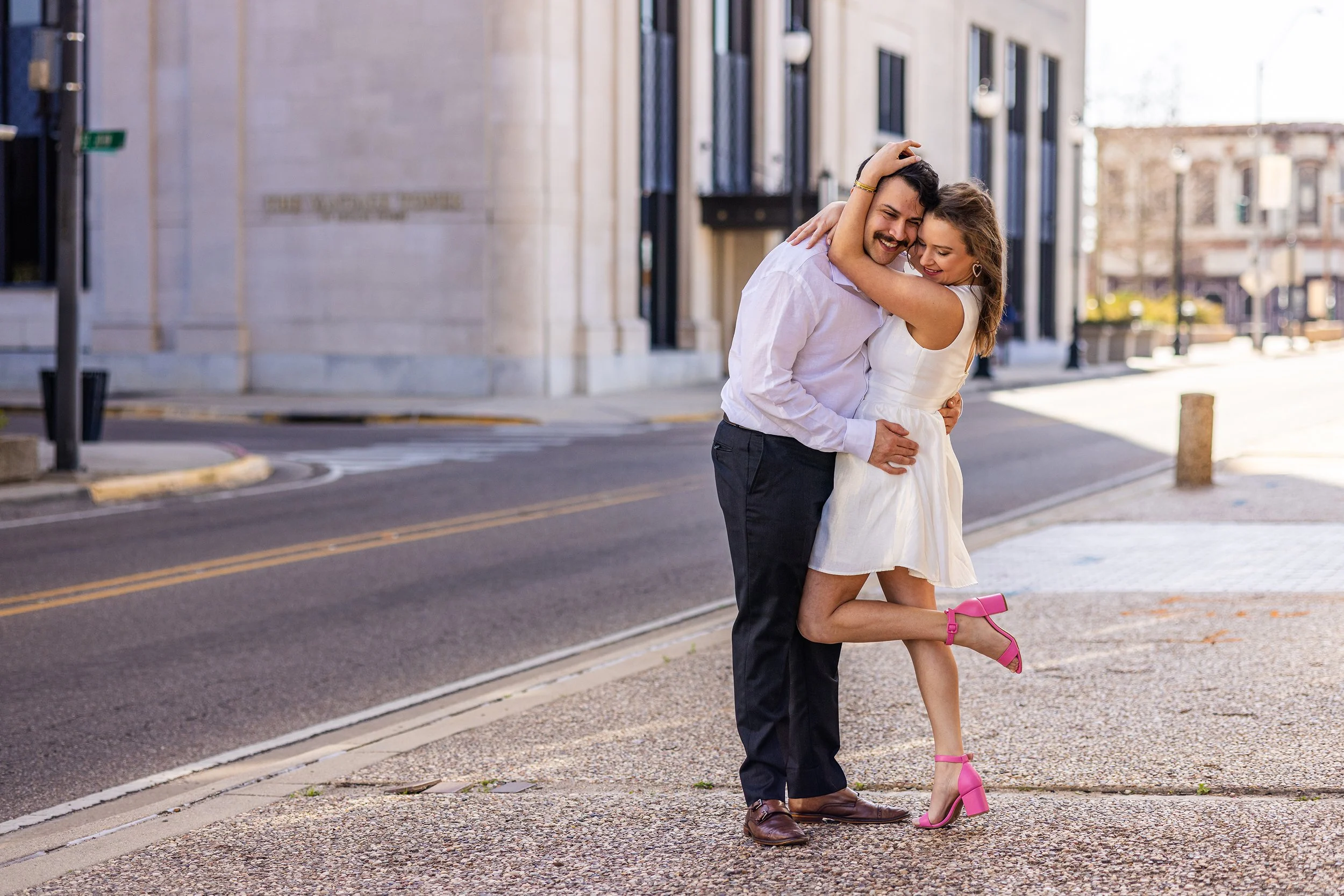 A happy couple embracing on a sidewalk in an urban area, with buildings and a street in the background. The woman is wearing a white dress and pink shoes; the man is in a white shirt and dark pants.