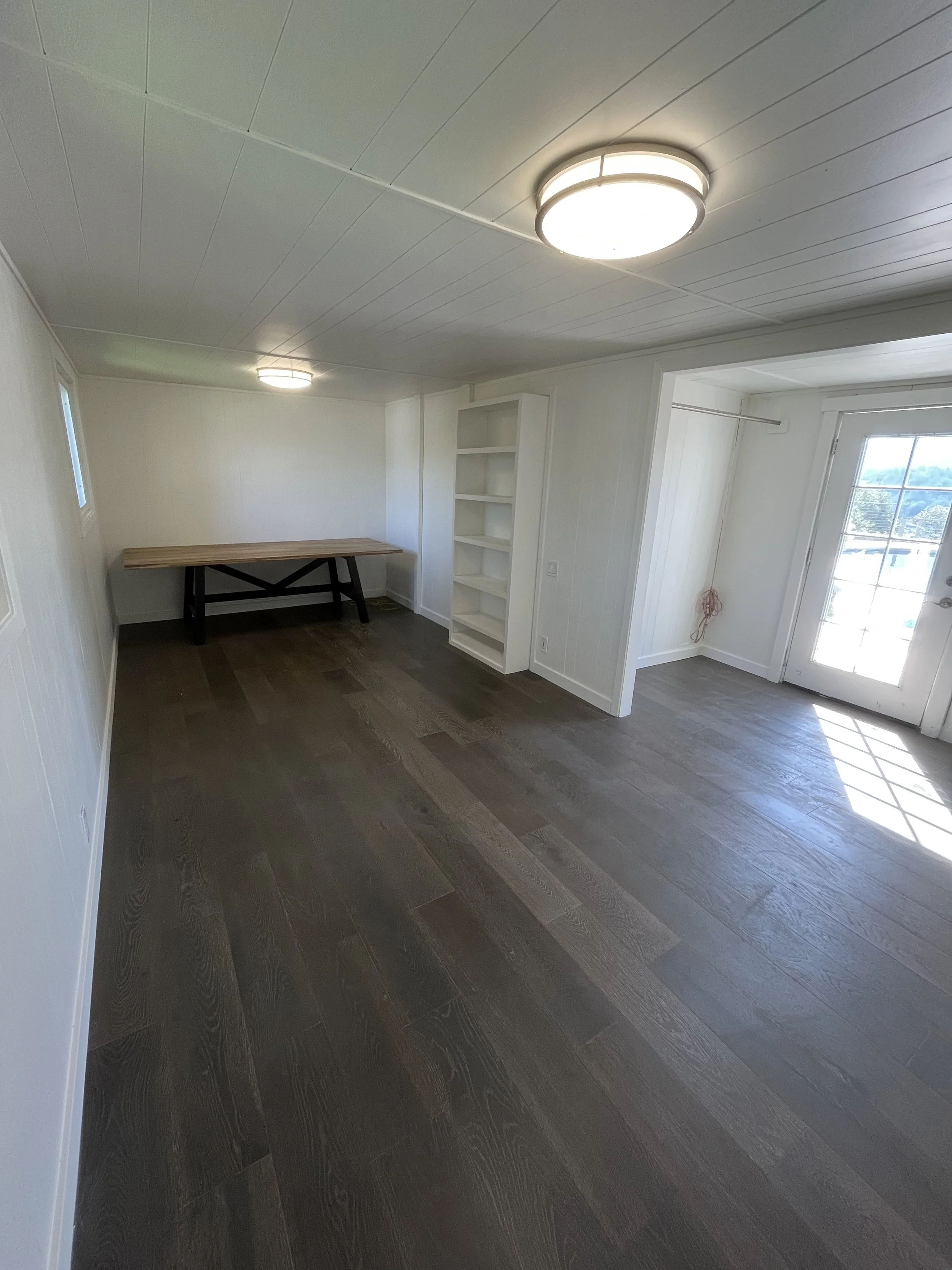 Empty room with dark hardwood floors, white paneled walls, ceiling lights, a window, a door with glass panes, and a wooden table in the corner.