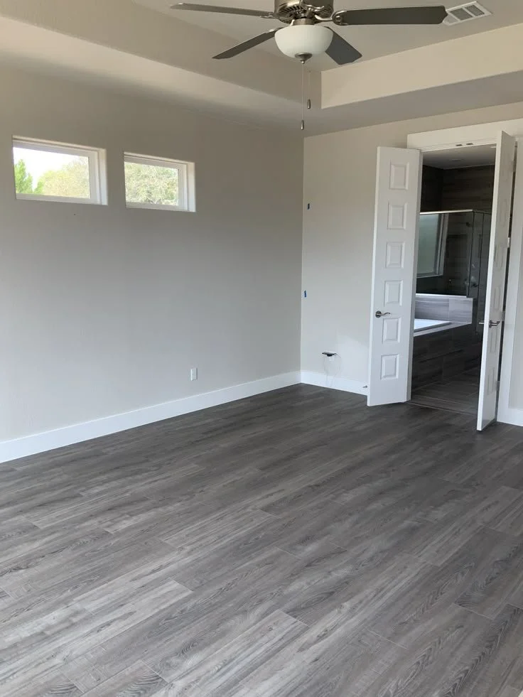 An empty room with light gray walls, hardwood flooring, a ceiling fan, three small windows, and an open door leading to a bathroom with a shower.