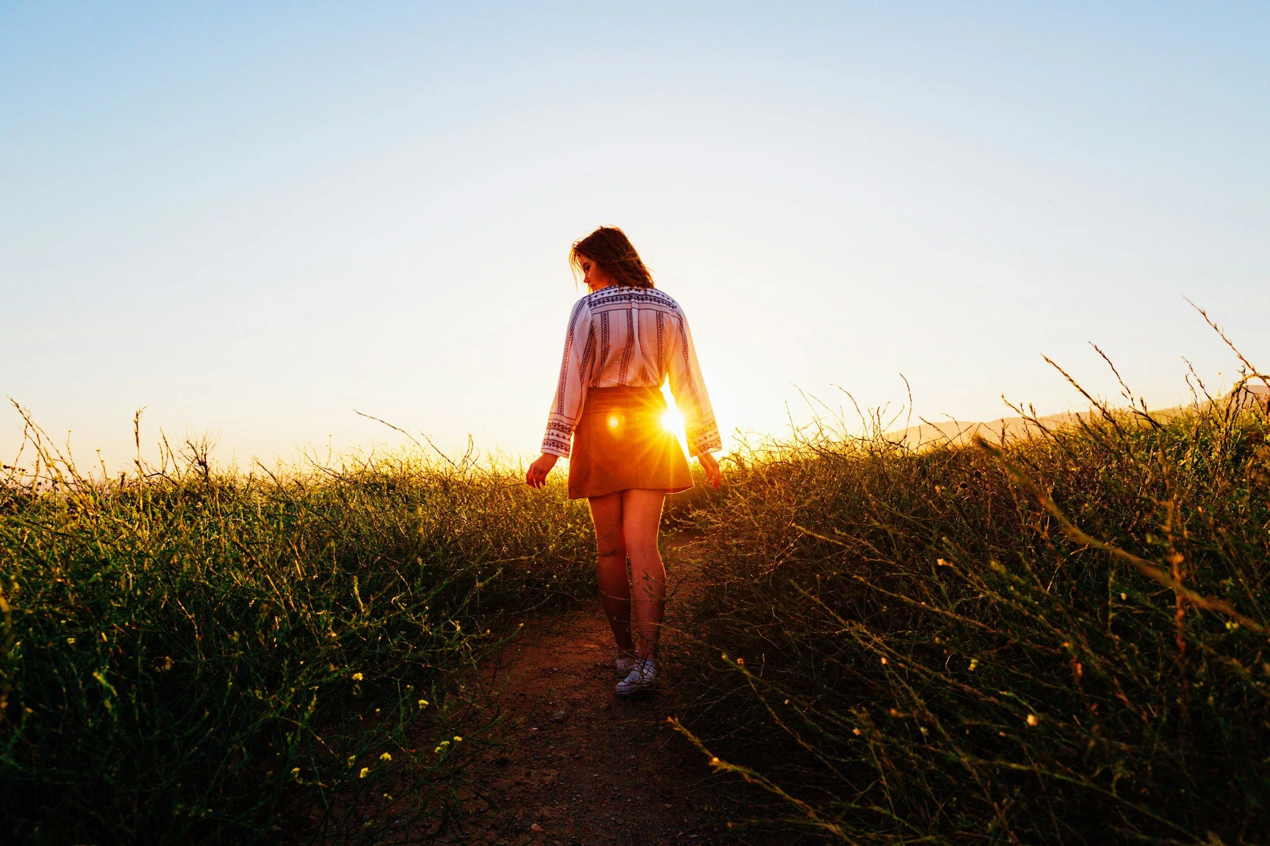 Woman walking on a dirt trail through tall grass during sunset or sunrise, with the sun shining directly behind her. On a journey, a path of discovery and connection with nature and essence.