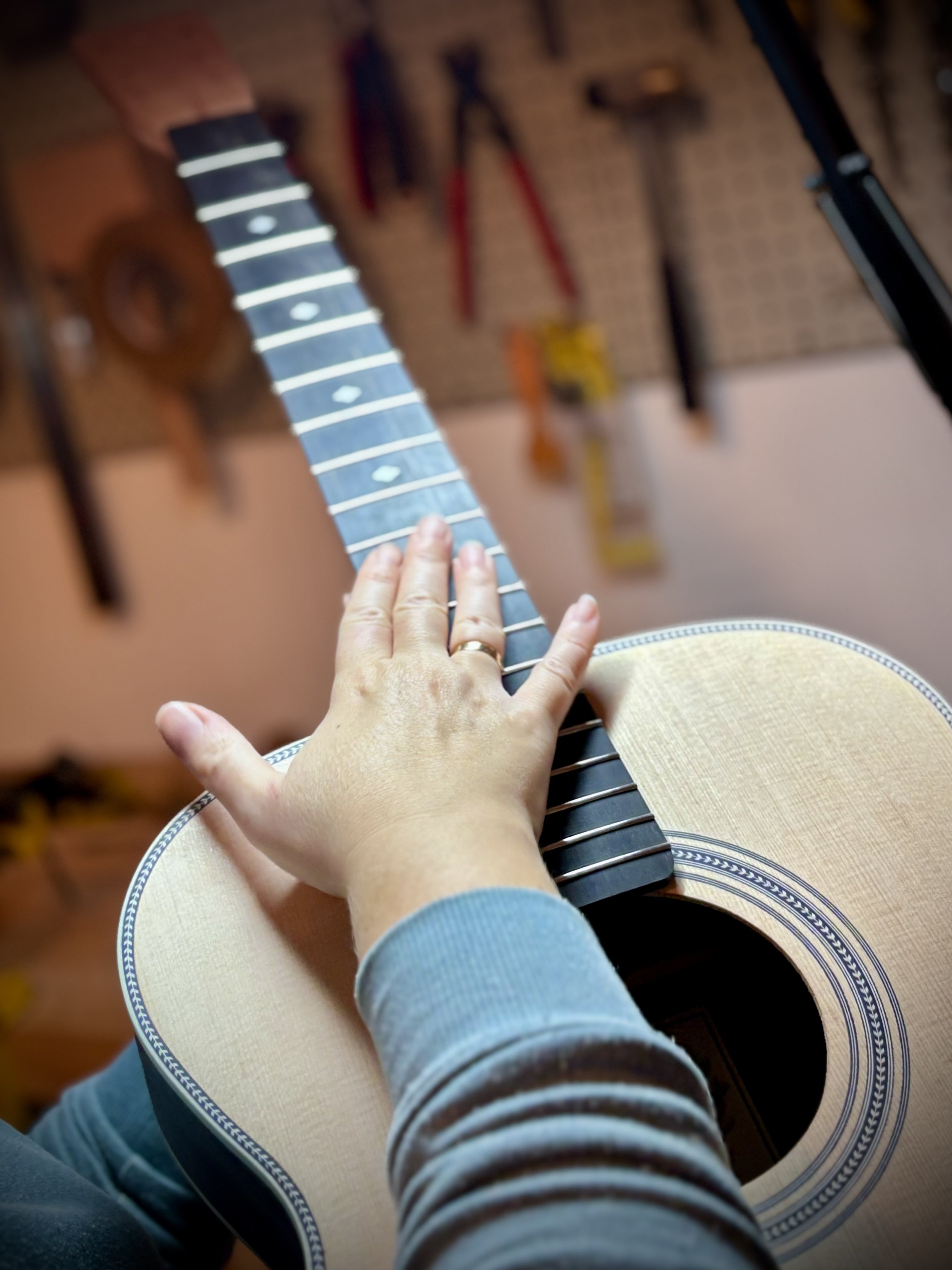 Una mano che suona su una chitarra acustica senza corde. Sullo sfondo, strumenti e utensili pendono da una parete di un laboratorio o di un'officina.