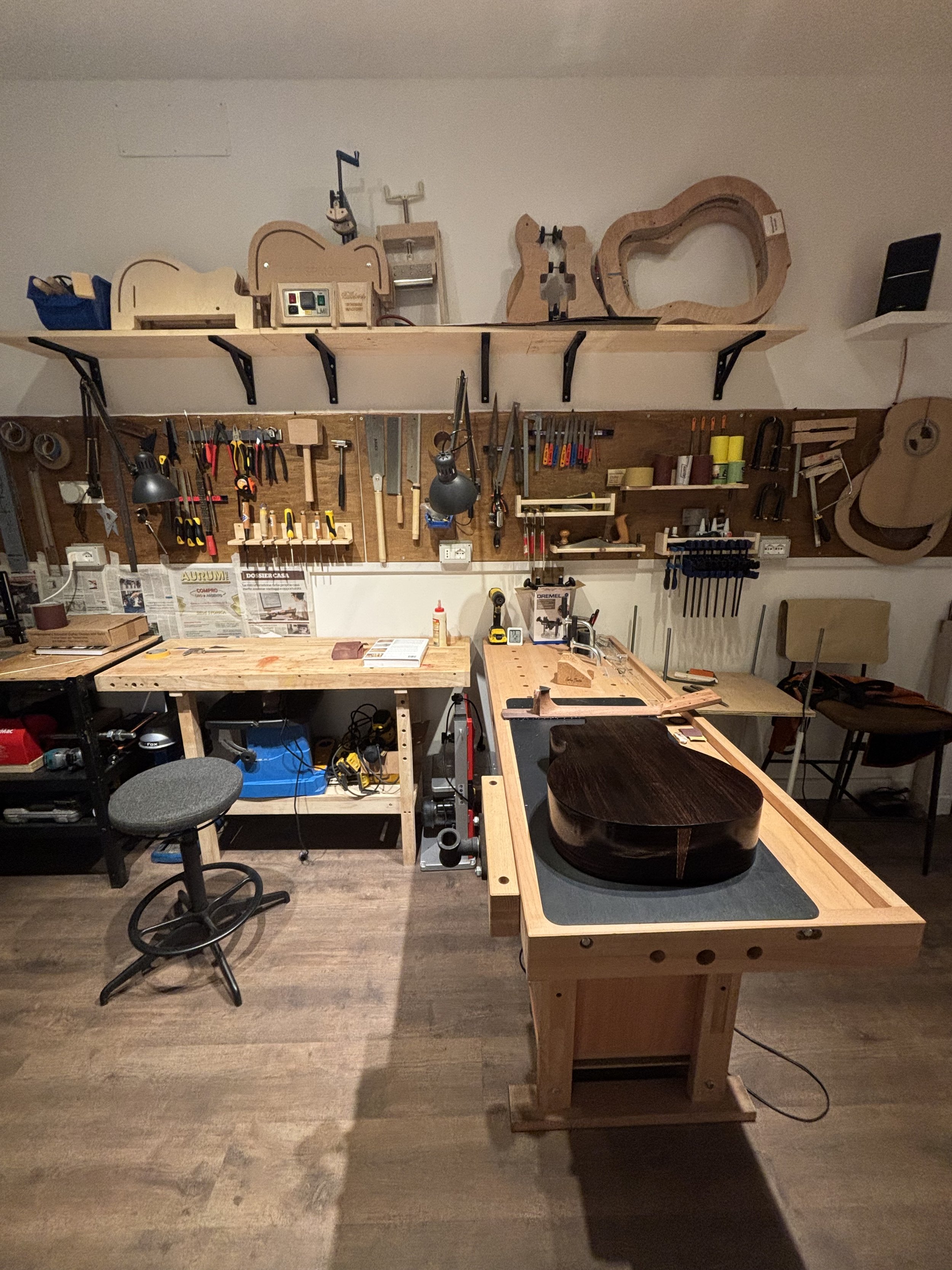 Laboratorio di falegnameria con strumenti e attrezzature per la lavorazione del legno, tra cui chitarre in fase di costruzione e strumenti da lavoro appesi alle pareti.