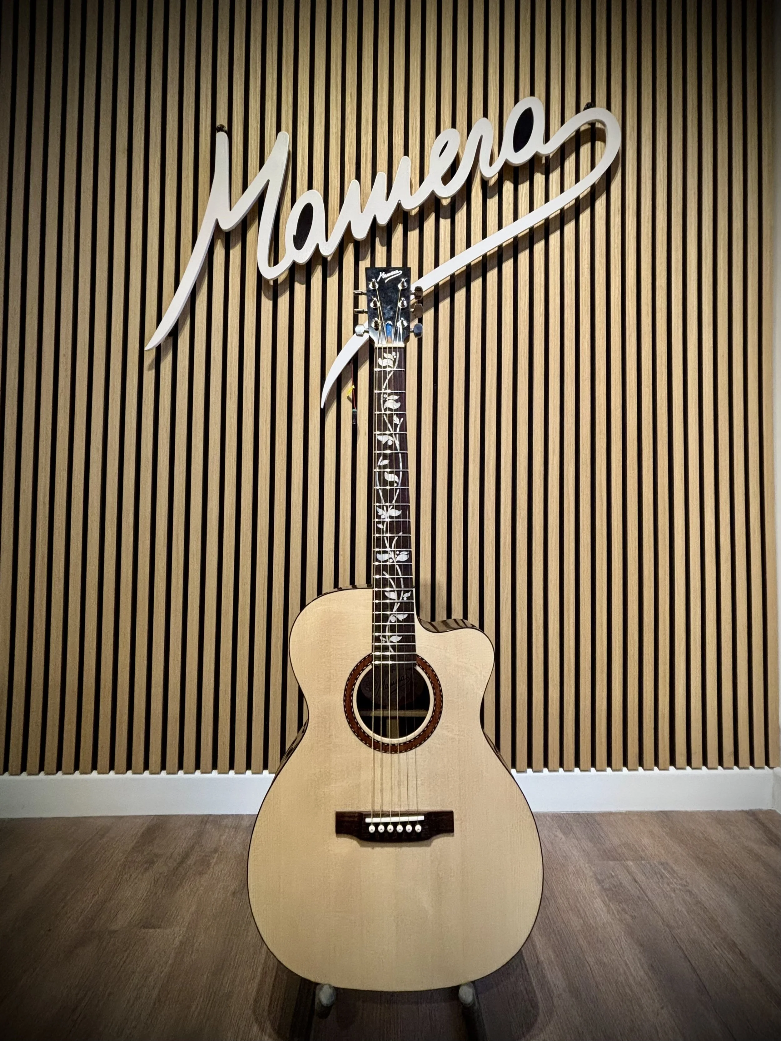 Chitarra acustica appoggiata a un support di fronte a una parete di pannelli di legno e sopra una scritta luminosa 'mamerla'.