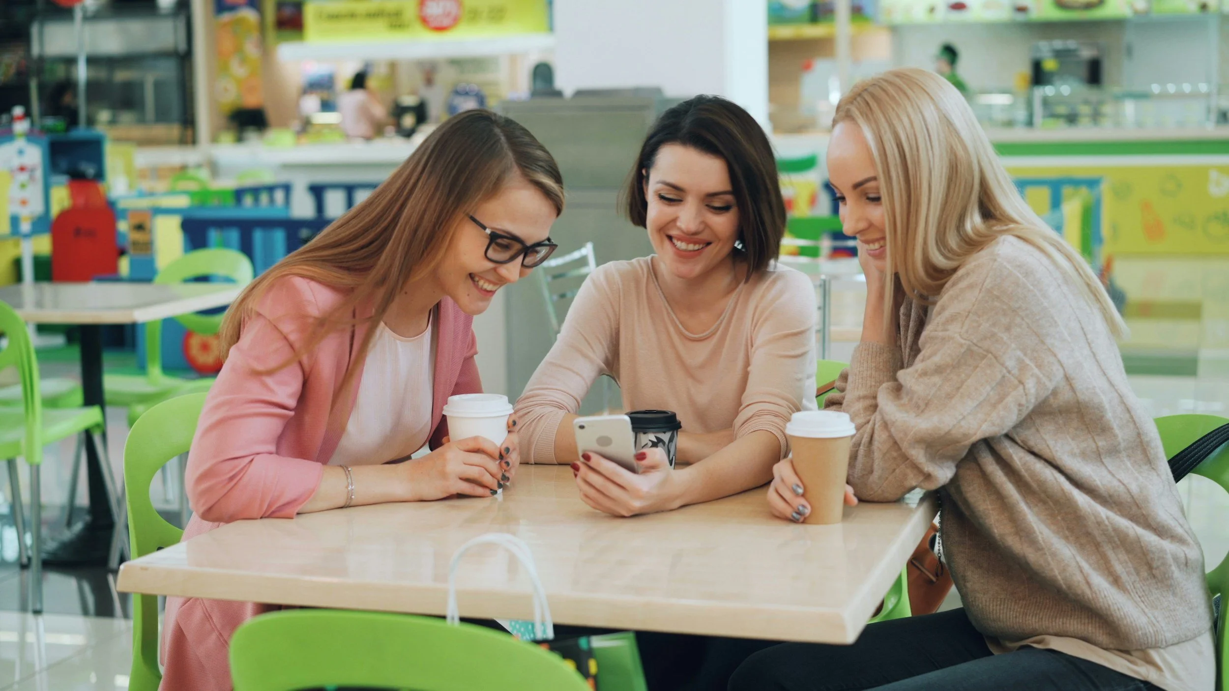 Three women sitting at a table in a brightly colored cafe, looking at a phone together and smiling, each holding a cup of coffee.