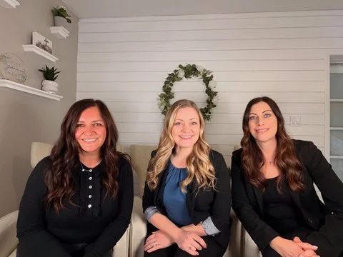 Three women sitting together indoors, smiling, with a white shiplap wall and greenery decoration behind them.
