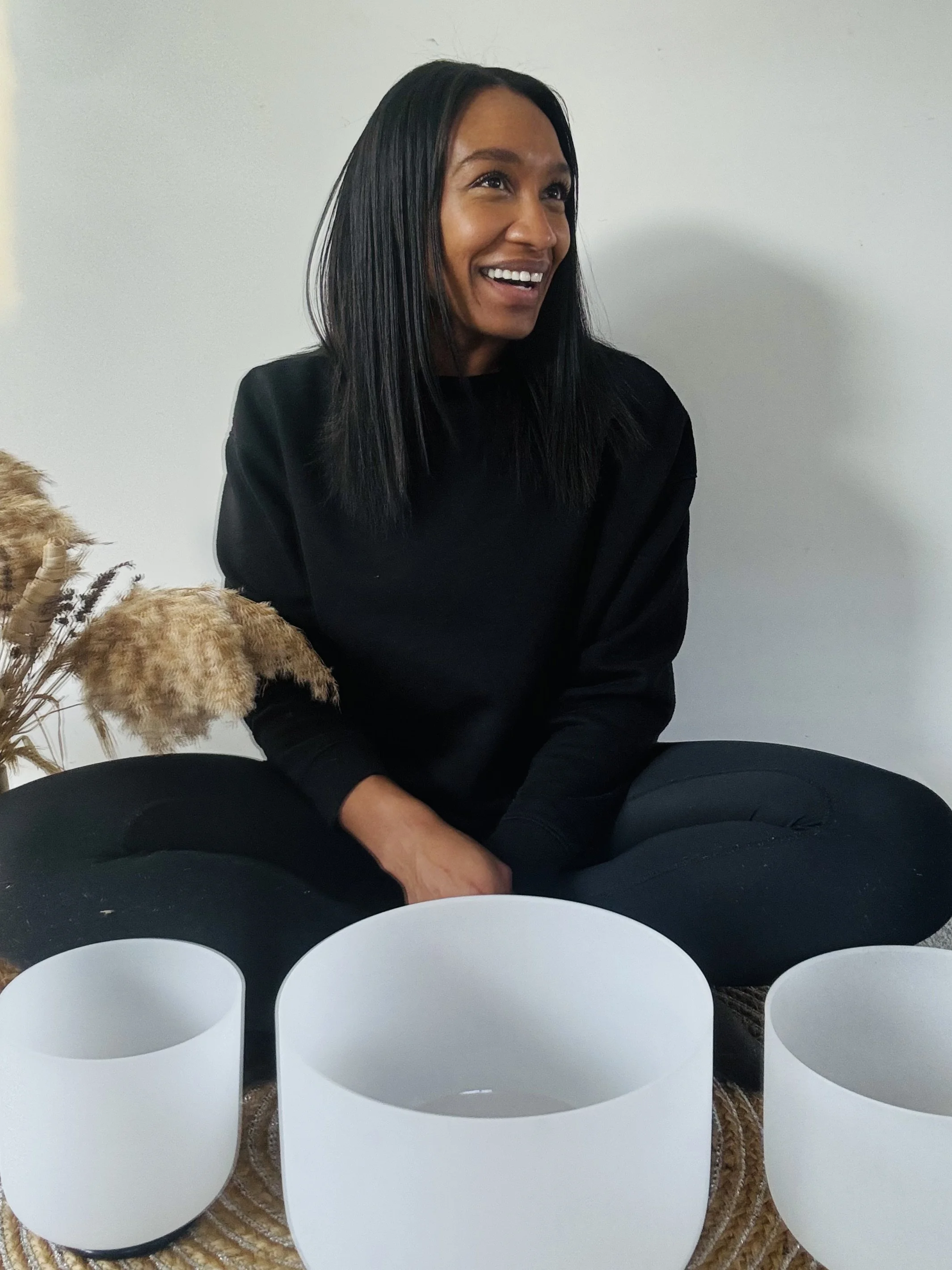 A woman sitting cross-legged on the floor, smiling, with three white singing bowls in front of her and decorative dried pampas grass to her left.