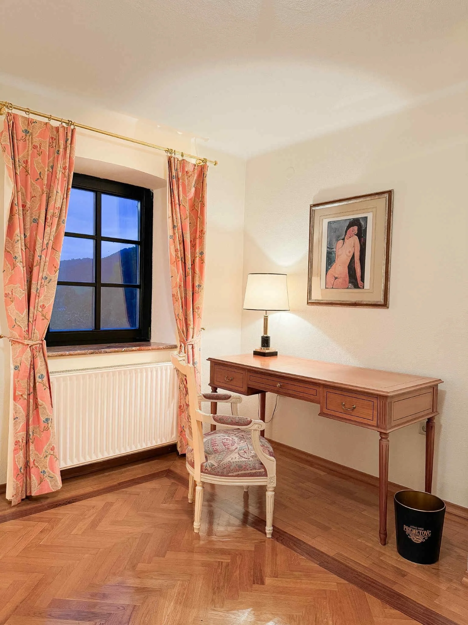A cozy room with a window, pink floral curtains, a wooden desk with a lamp, a decorative chair, a wall painting of a woman, and a black wastebasket on a wooden floor.