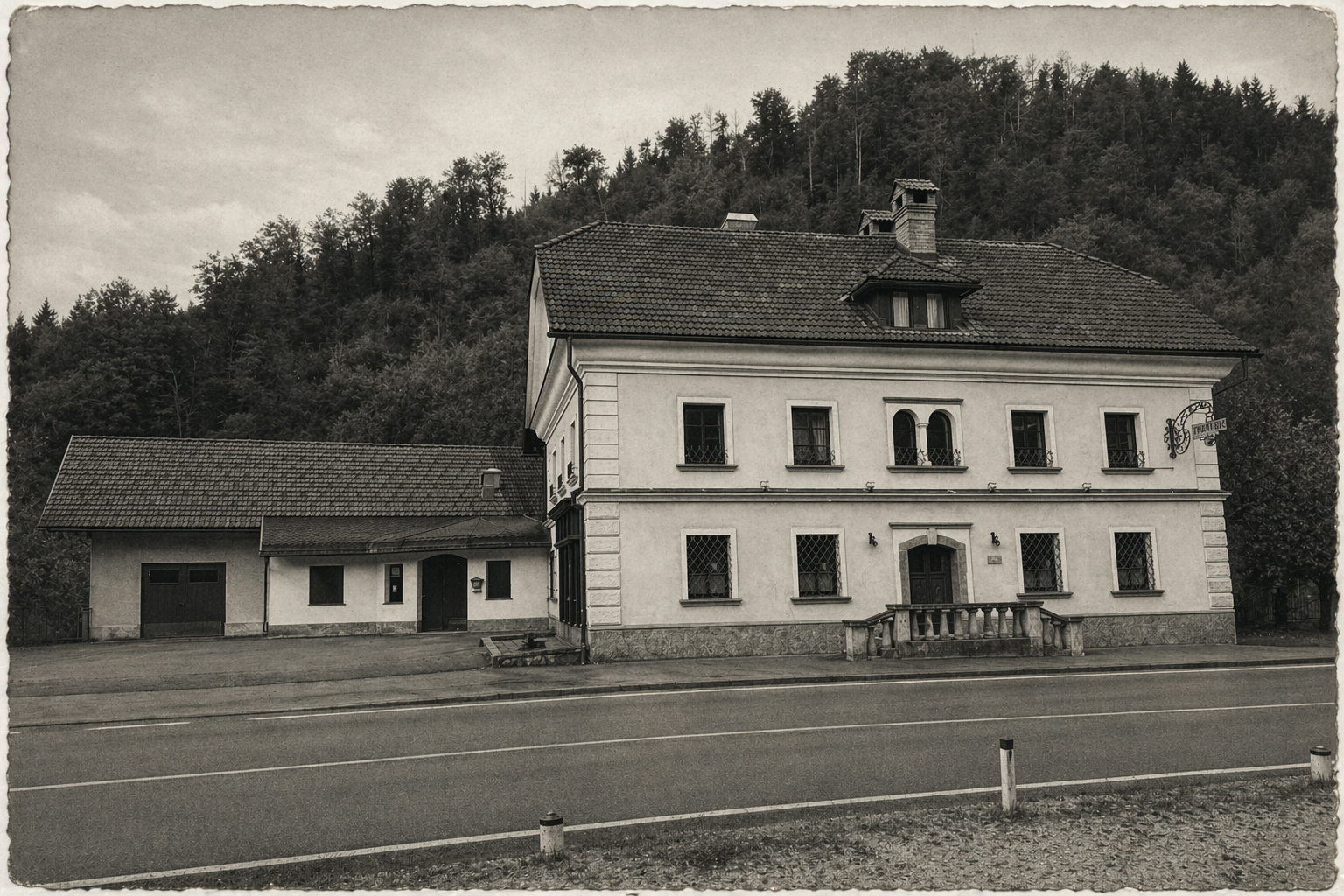 Black and white photo of The Premetovc with a small attached garage, surrounded by trees and hills, with a road in front.