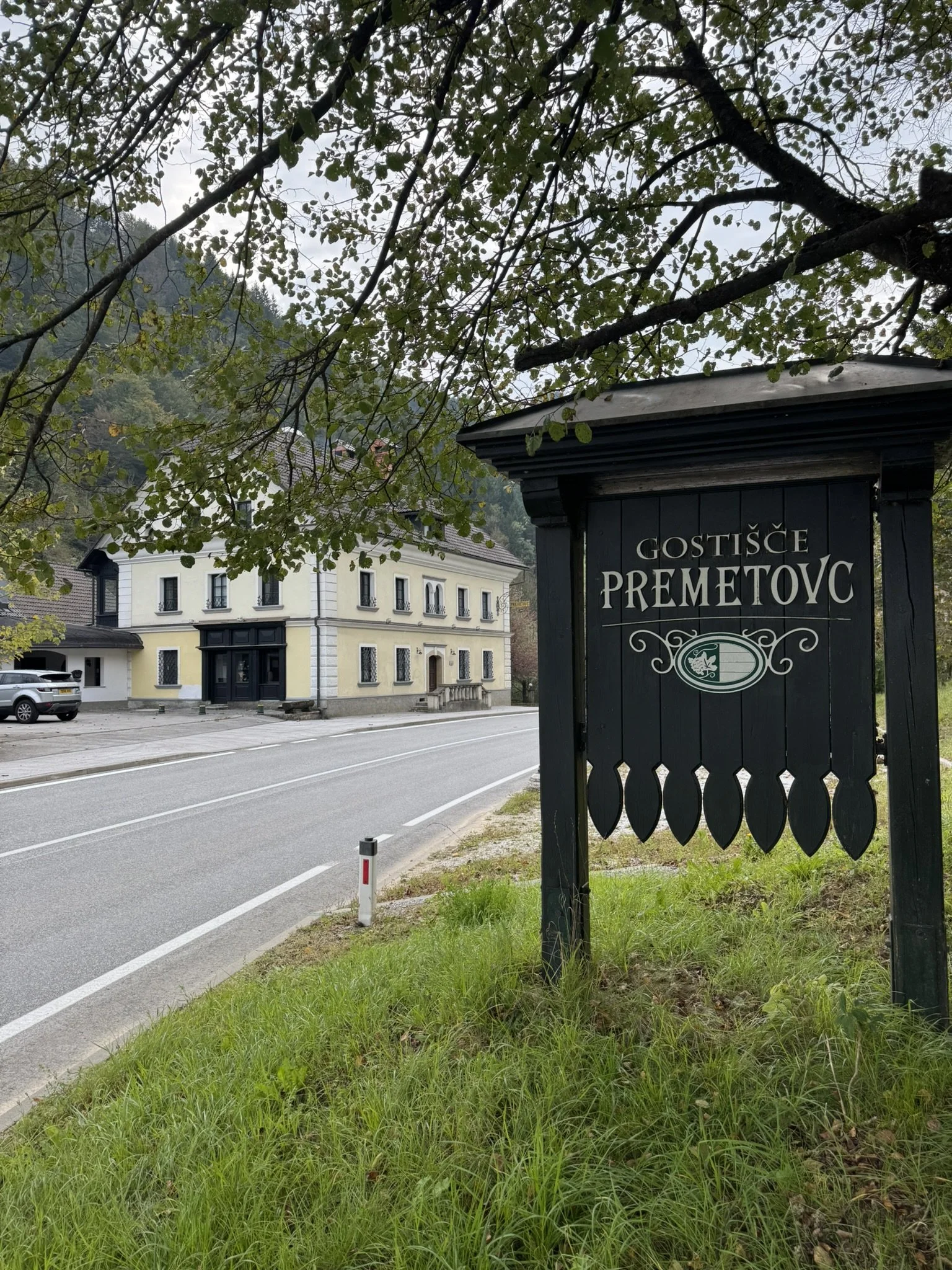 A street-side sign reading 'GOSTIŠČE PREMOTOV' in front of a white building with black accents, next to a road with parked cars and a hillside with trees in the background, under an overcast sky.