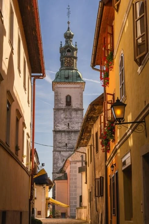 A narrow European street lined with colorful buildings and flower pots, with a tall church tower with a green dome and clock in the background.