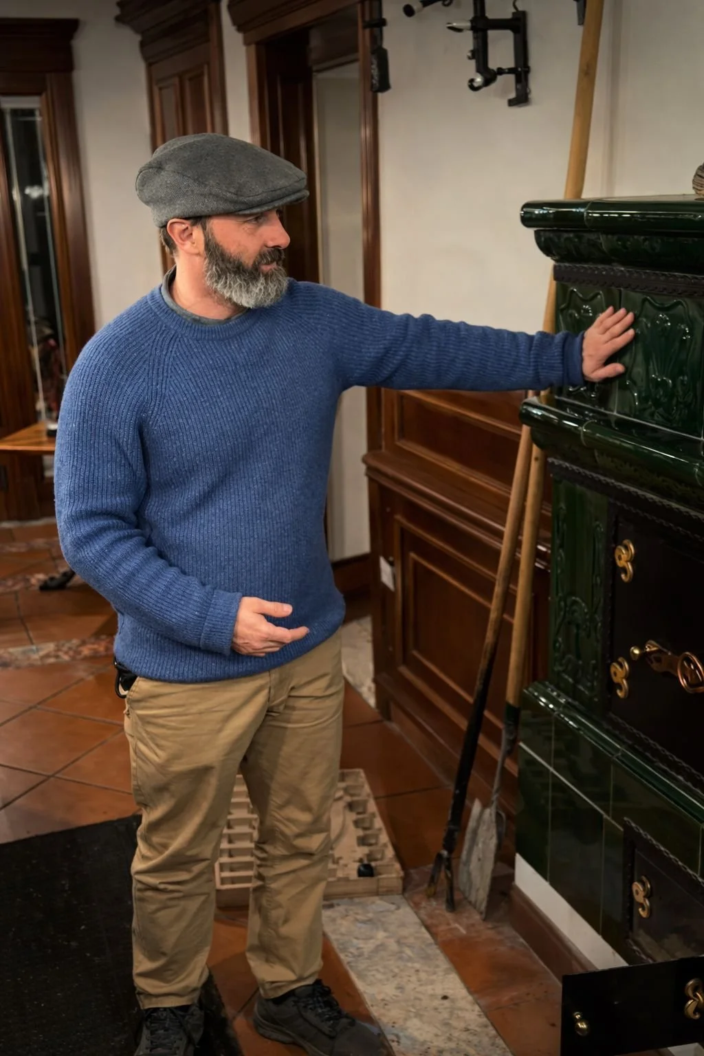 A man with a gray beard, wearing a gray flat cap and a blue sweater, stands indoors, touching a dark green ornate ceramic stove or oven.