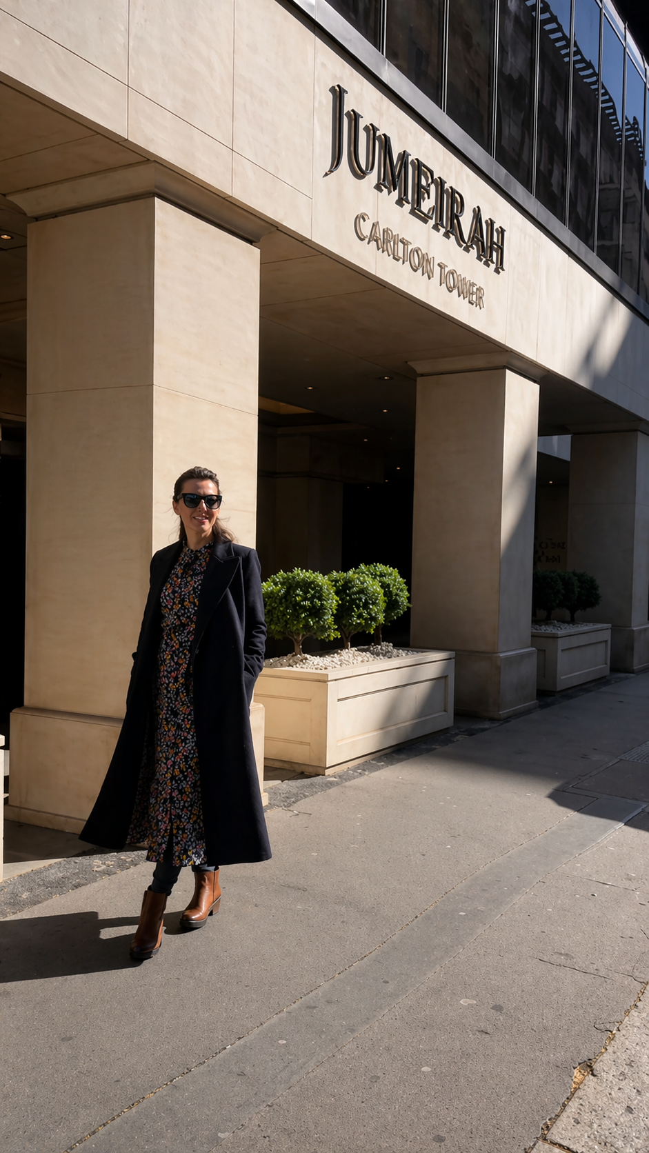 A woman in a black coat and sunglasses standing outside of Jumeirah Carlton Tower hotel in London on a sunny day.