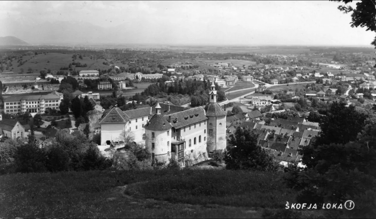 Black and white aerial view of a historical castle with towers, surrounded by a town with various buildings and houses, and open landscapes in the background. The text at the bottom right reads "Škofja Loka."