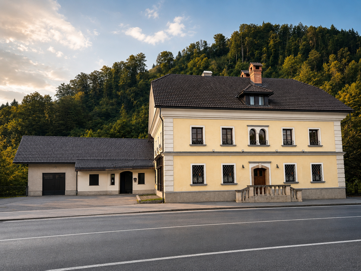A two-story yellow house with a dark roof, situated at the edge of a road, with green forested hills in the background.