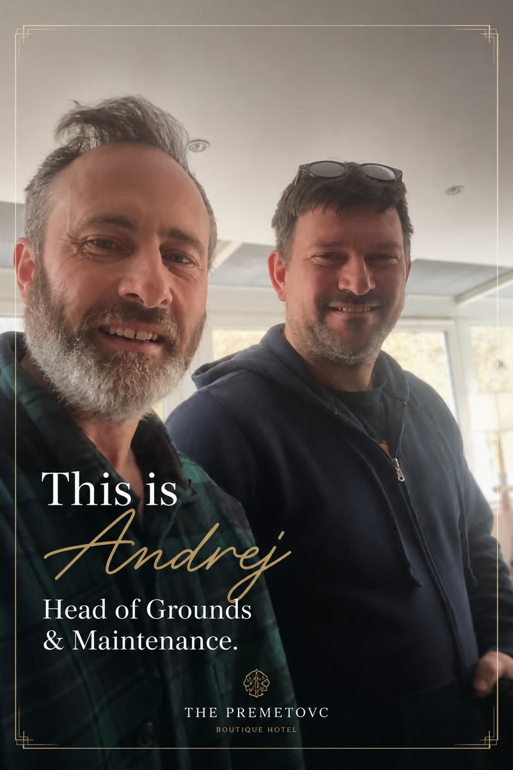 Two men smiling for a photo indoors, one with a beard and gray hair wearing a checkered shirt, and the other with glasses on his head wearing a dark hoodie.