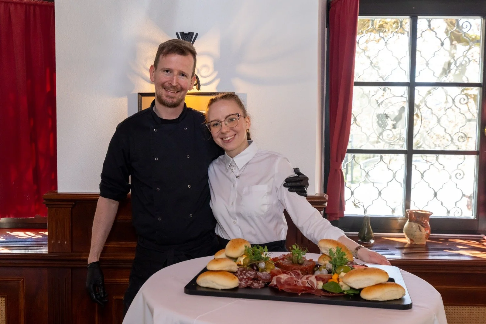 A man and woman standing behind a table with a charcuterie and bread platter. The man is wearing a black chef's coat, and the woman is wearing glasses and a white shirt. They are smiling and posing for the photo inside a room with large windows and red curtains.