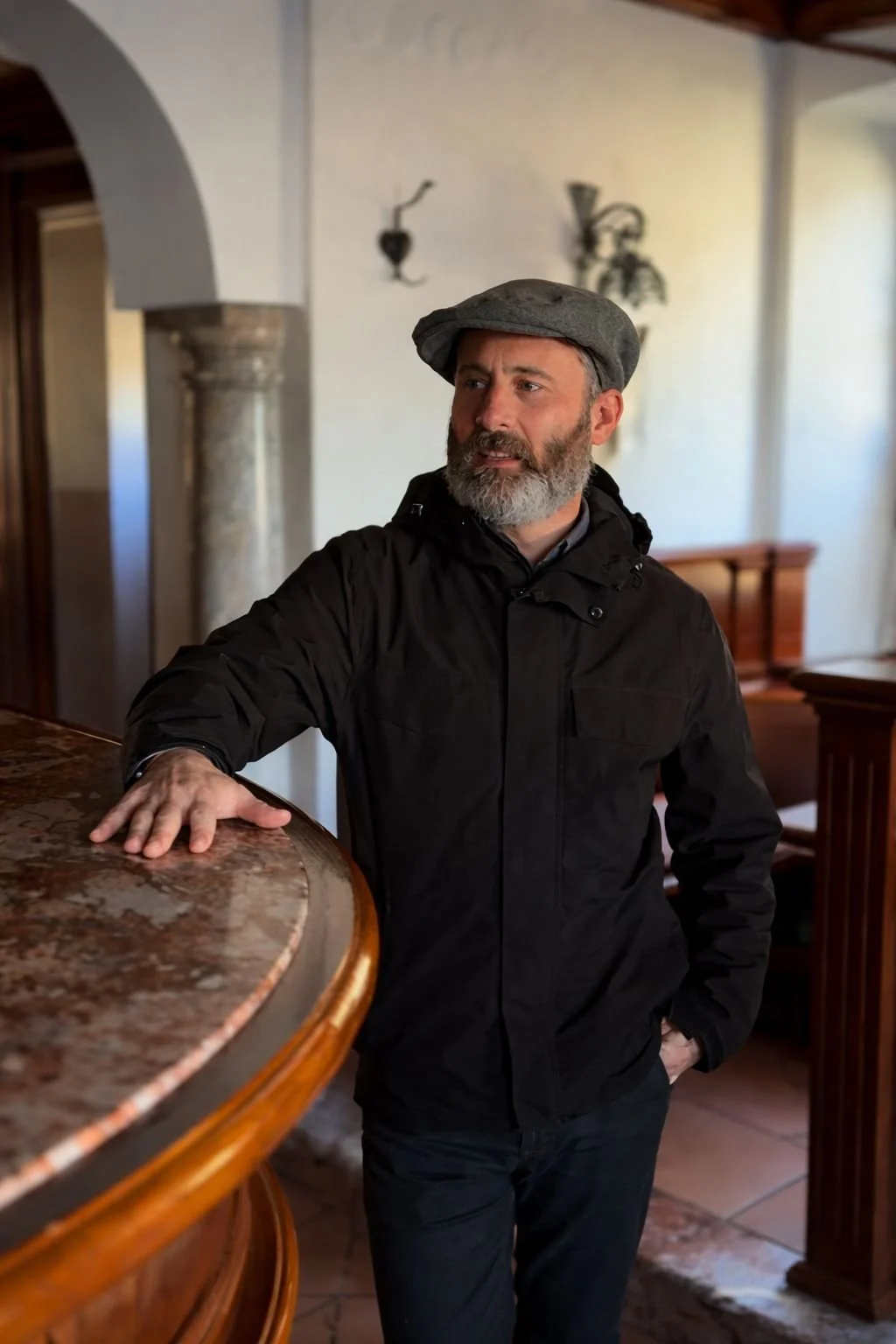 A man with a beard and gray hair wearing a gray flat cap and black jacket, standing indoors, resting his hand on a marble countertop, looking to the side.