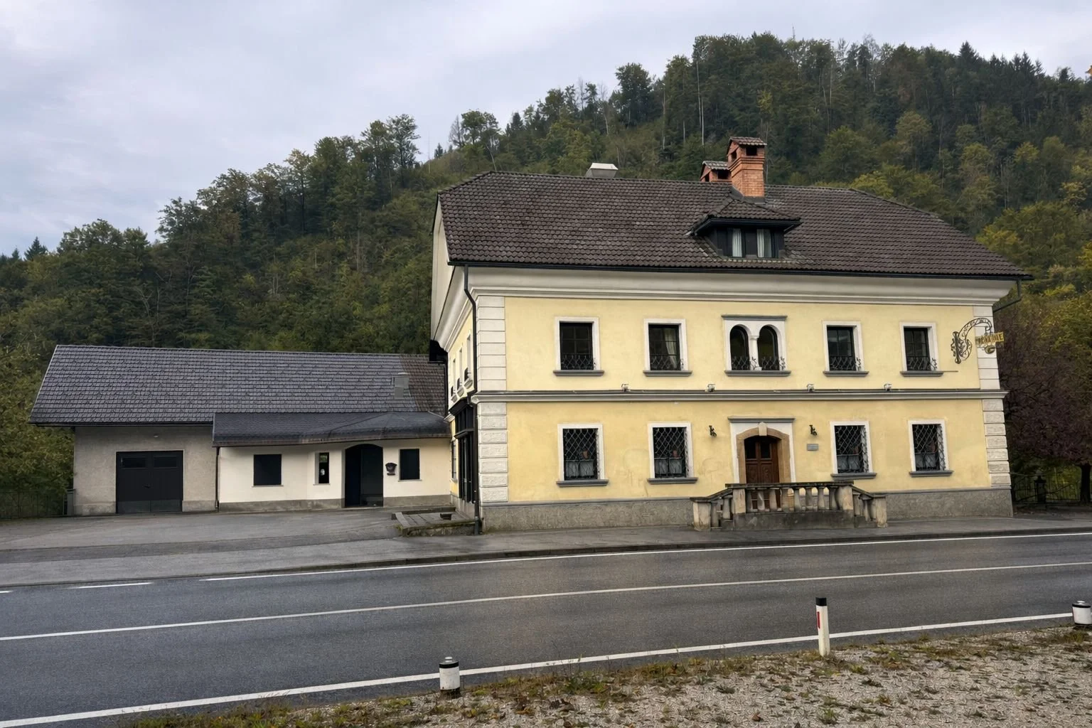 A two-story yellow house with a brown roof, surrounded by a mountainous landscape with trees, next to a smaller white building, along a paved road.