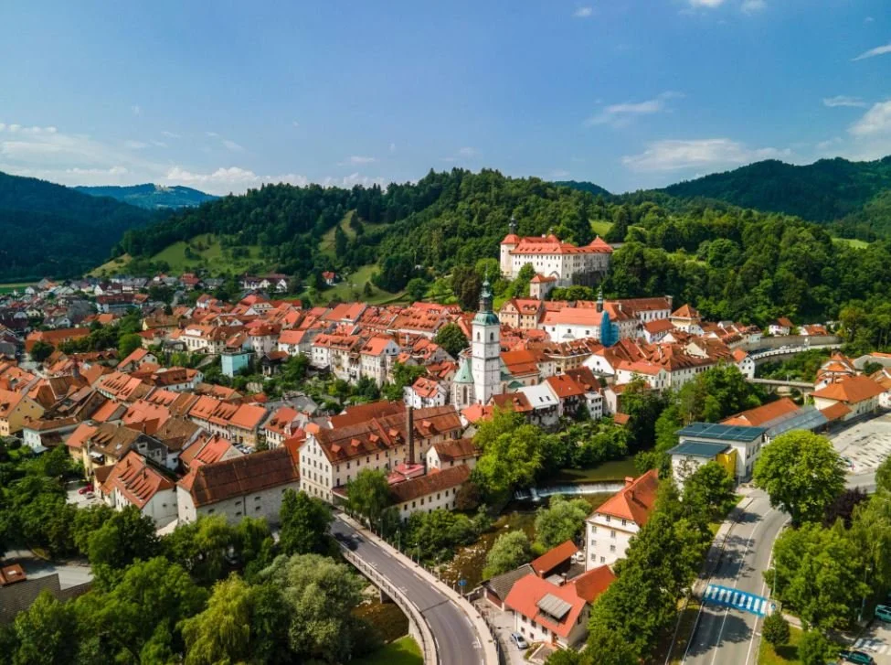 A picturesque town surrounded by green hills and mountains, featuring red-roofed buildings, a prominent church with twin steeples, and a historic castle on the hilltop under a blue sky.