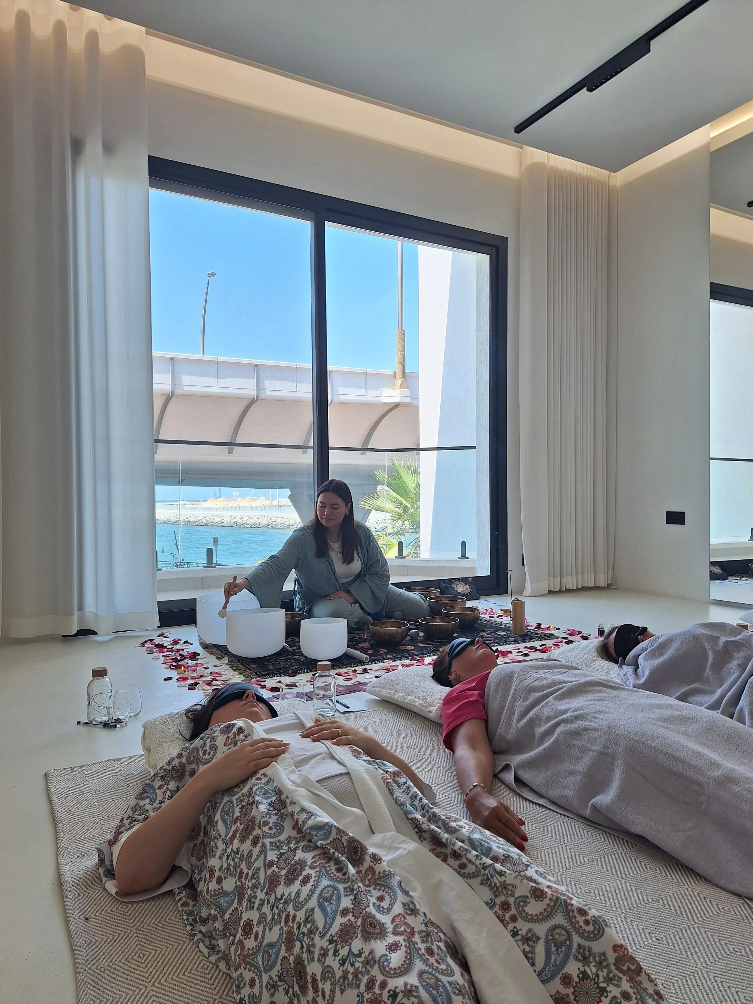 People lying on mats with headphones on during a sound healing session in a room with large windows and a view of water outside.