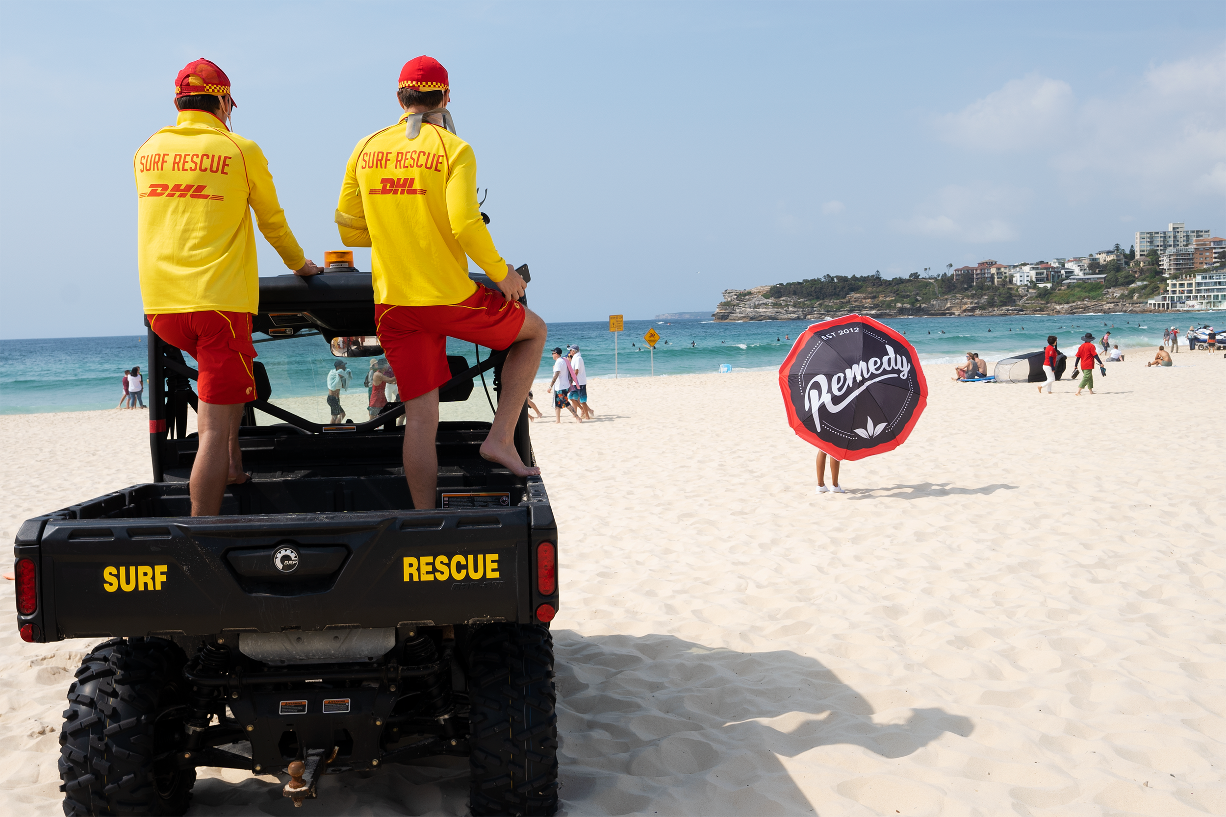 Two surf rescue personnel in yellow shirts and red shorts on a rescue vehicle at a beach. One person is sitting, and the other is standing, both wearing red hats. The beach has many visitors, with some using umbrellas and the ocean in the background.