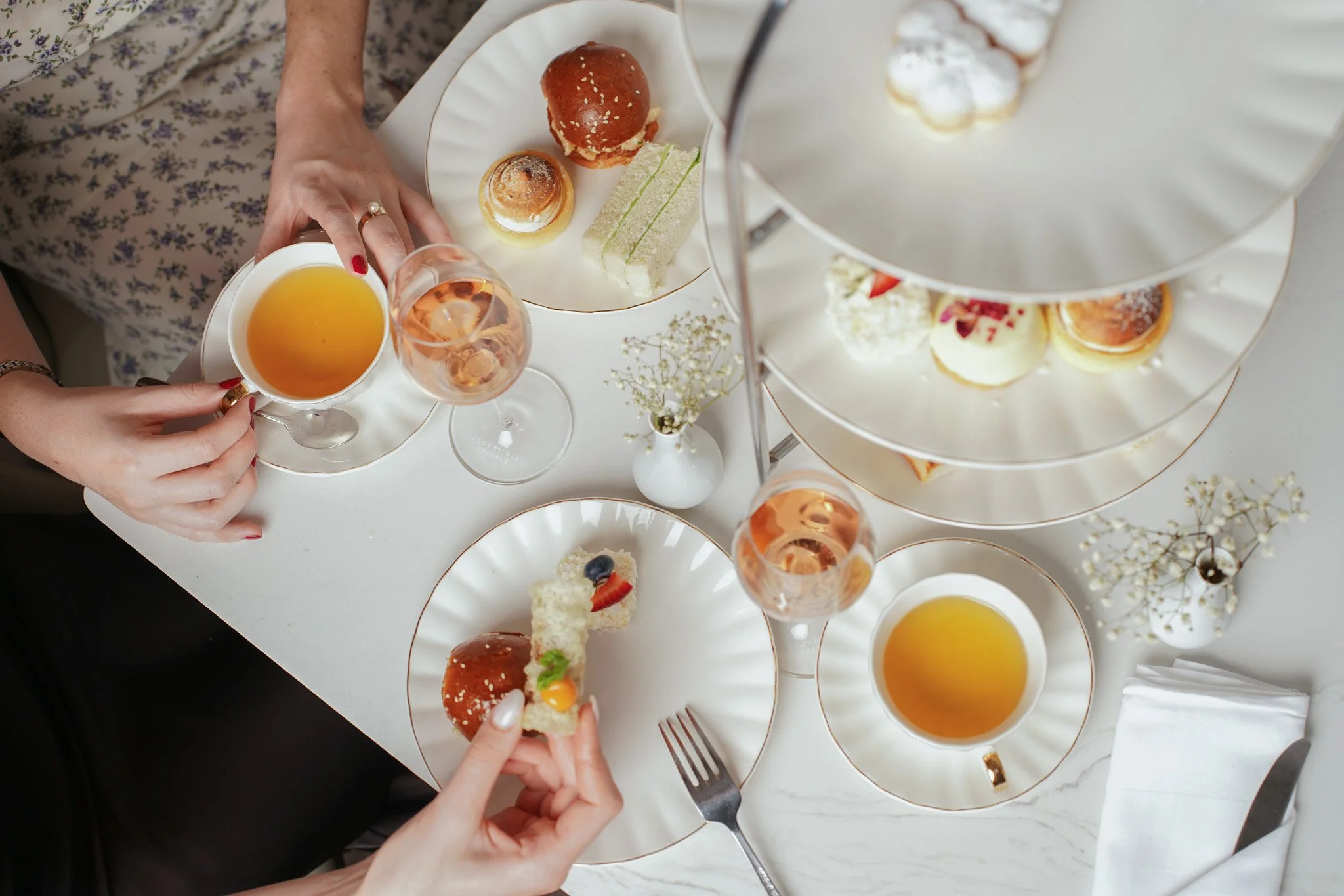 High tea editorial photography at Paradox Hotel Sydney - Two people holding small sandwiches at a tea party with tiered trays of assorted pastries, desserts, and drinks on a white table.