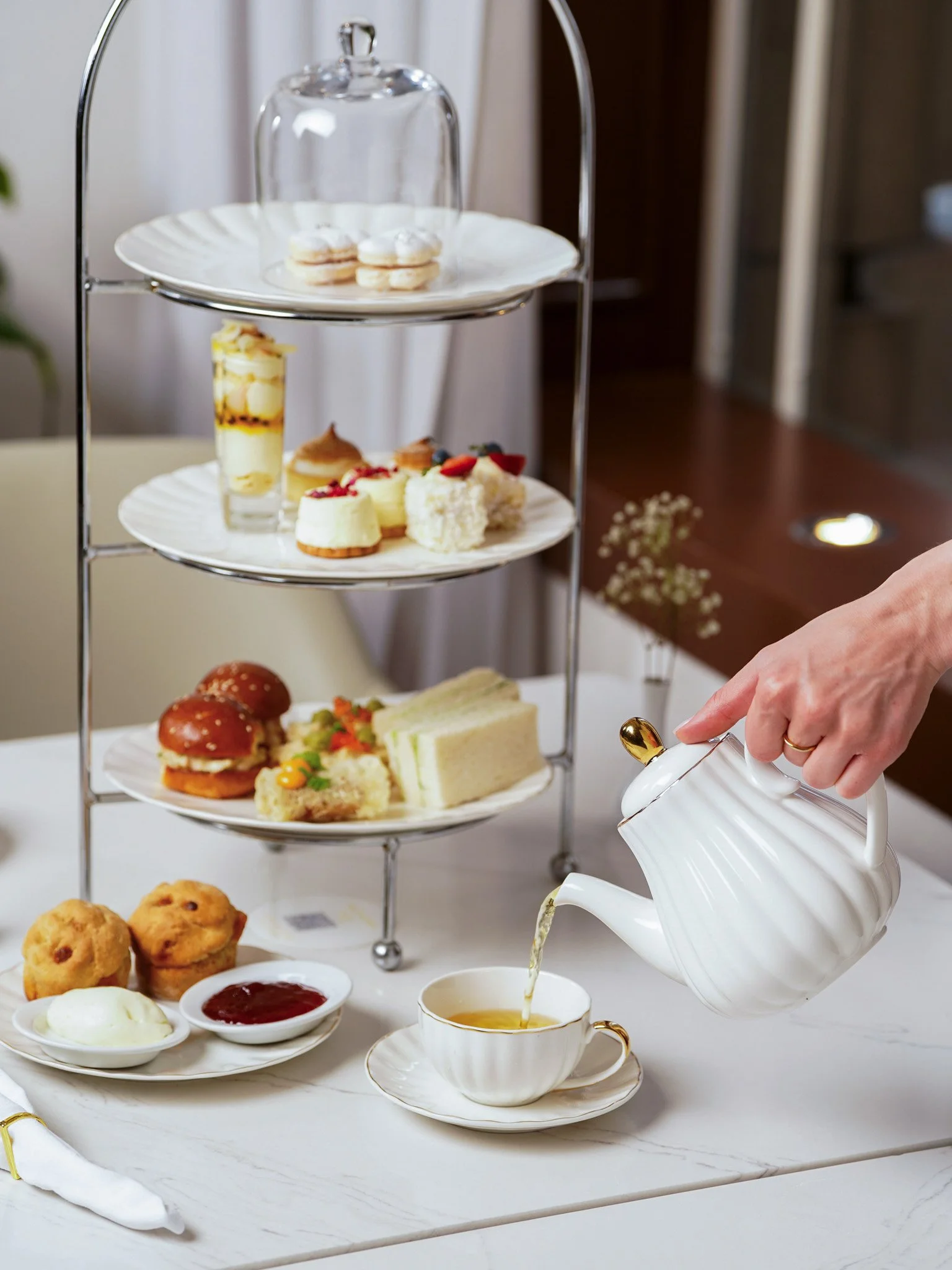 High tea food photography at Paradox Hotel Sydney - A three-tiered serving stand filled with various desserts and finger foods, with a hand pouring tea from a porcelain teapot into a teacup on a white table.