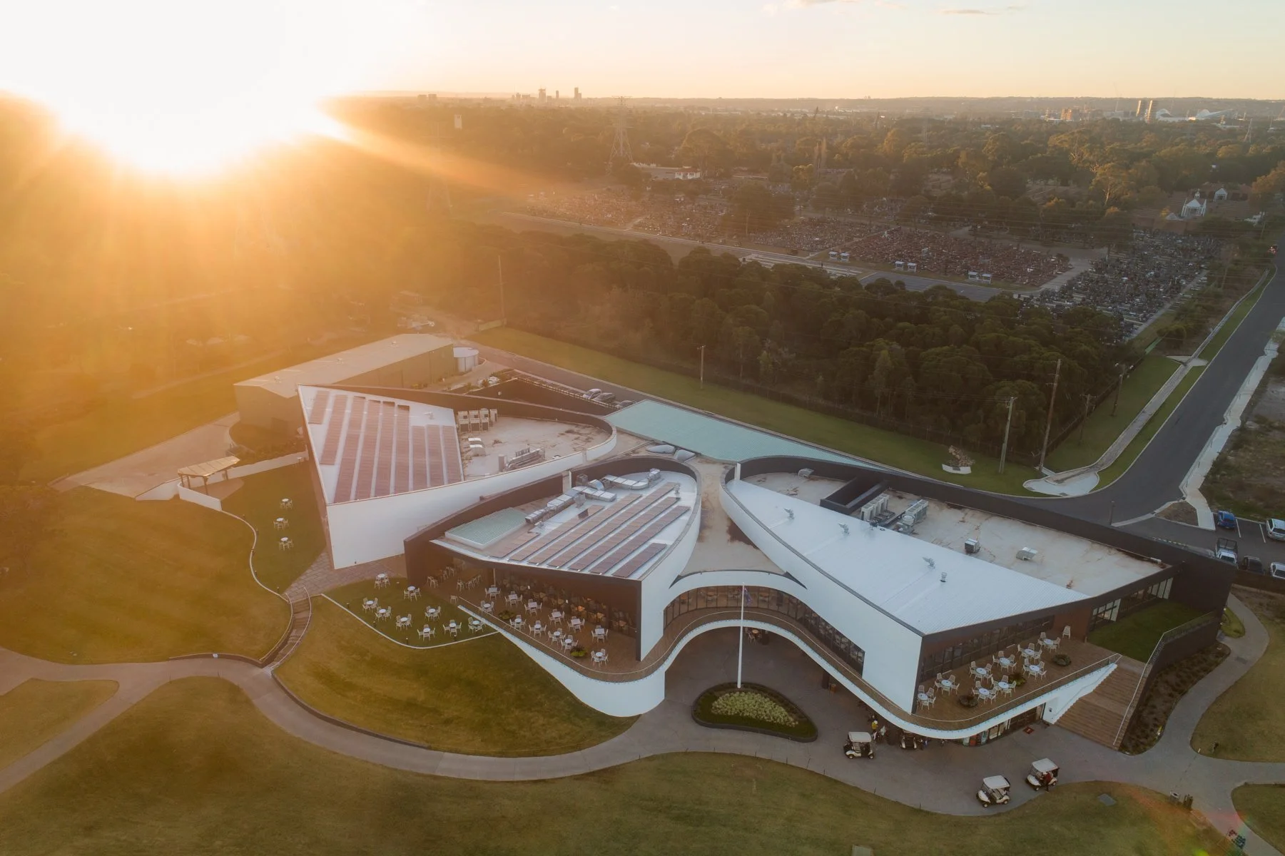 An aerial view of a modern building with a curved white roof, surrounded by green lawns and outdoor seating areas, with sunlight shining from the upper left corner and a city skyline in the distance.