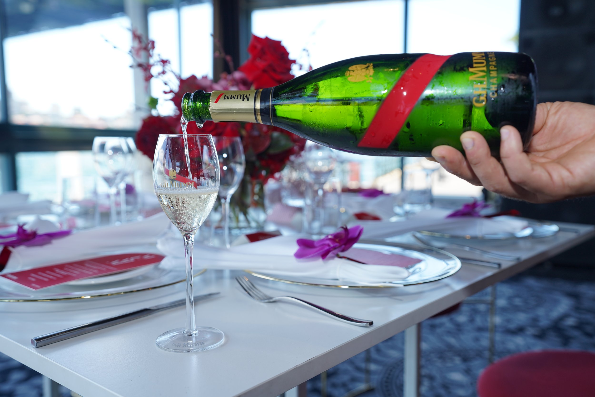 A person pouring champagne from a green bottle into a glass on a table decorated with pink flower petals, with a blurred background of windows and floral arrangements.
