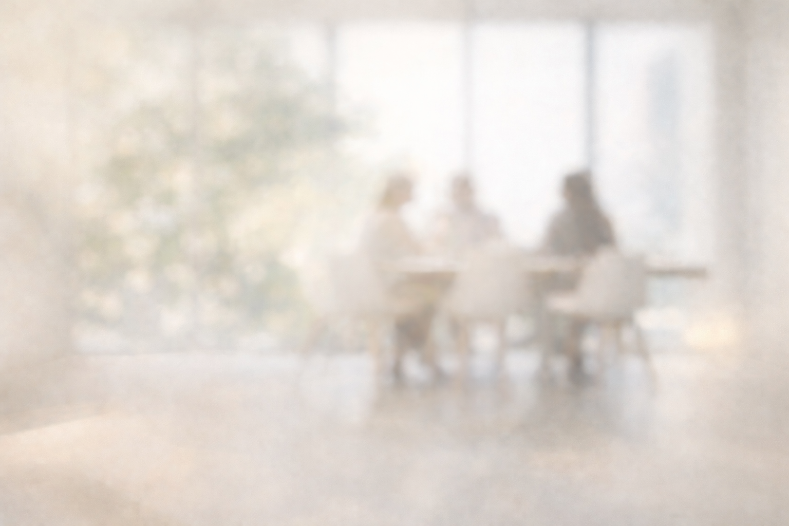 Three people sitting around a table in a bright room with large windows, engaged in a discussion or meeting.