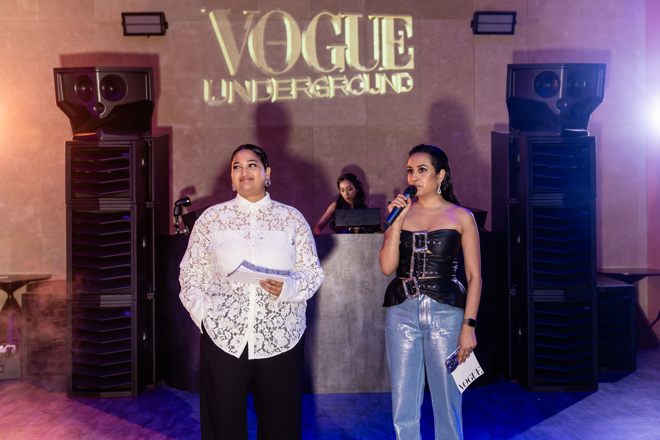 Two women speaking at Vogue Underground event with a DJ in the background, large speakers on either side, colorful lighting, and a projected Vogue Underground logo on the wall.