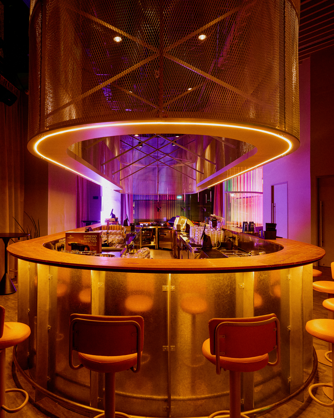 A modern, illuminated bar area with a curved wooden counter, golden metallic panels, and hanging pink and purple neon lights.