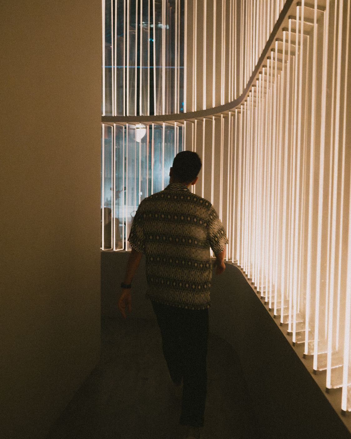 A person walking in a modern, dimly lit hallway with vertical white slats on the right side and an illuminated glass wall on the left side.