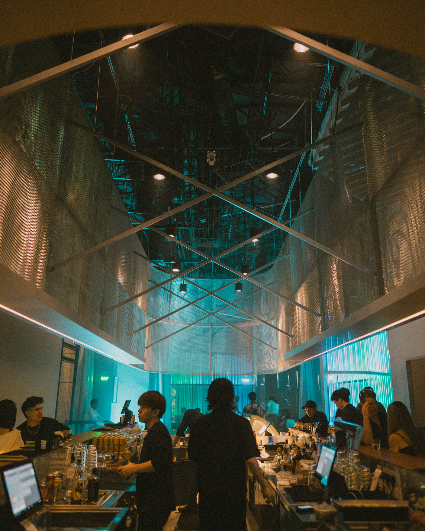 Interior of a modern bar or nightclub with a high, reflective ceiling with geometric metal framework, blue accent lighting, and people socializing at the bar.